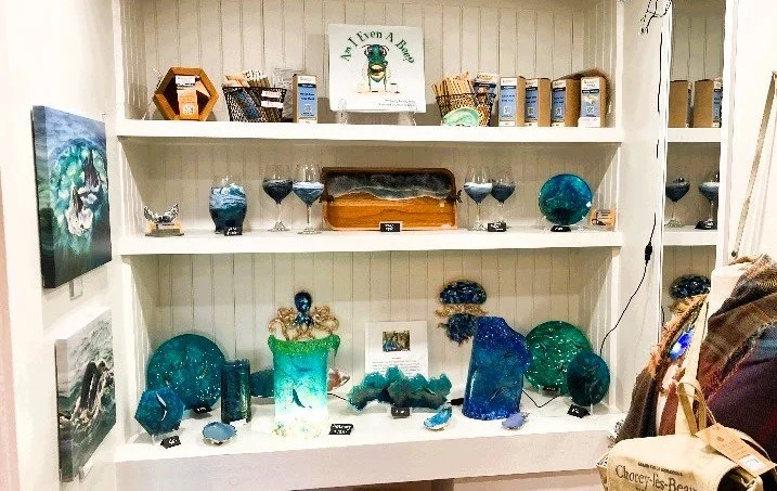 Display shelf with glass art and decor items, including blue and green glass pieces, vases, and sculptures. A small sign on the top shelf reads 'A Event A Br...'.