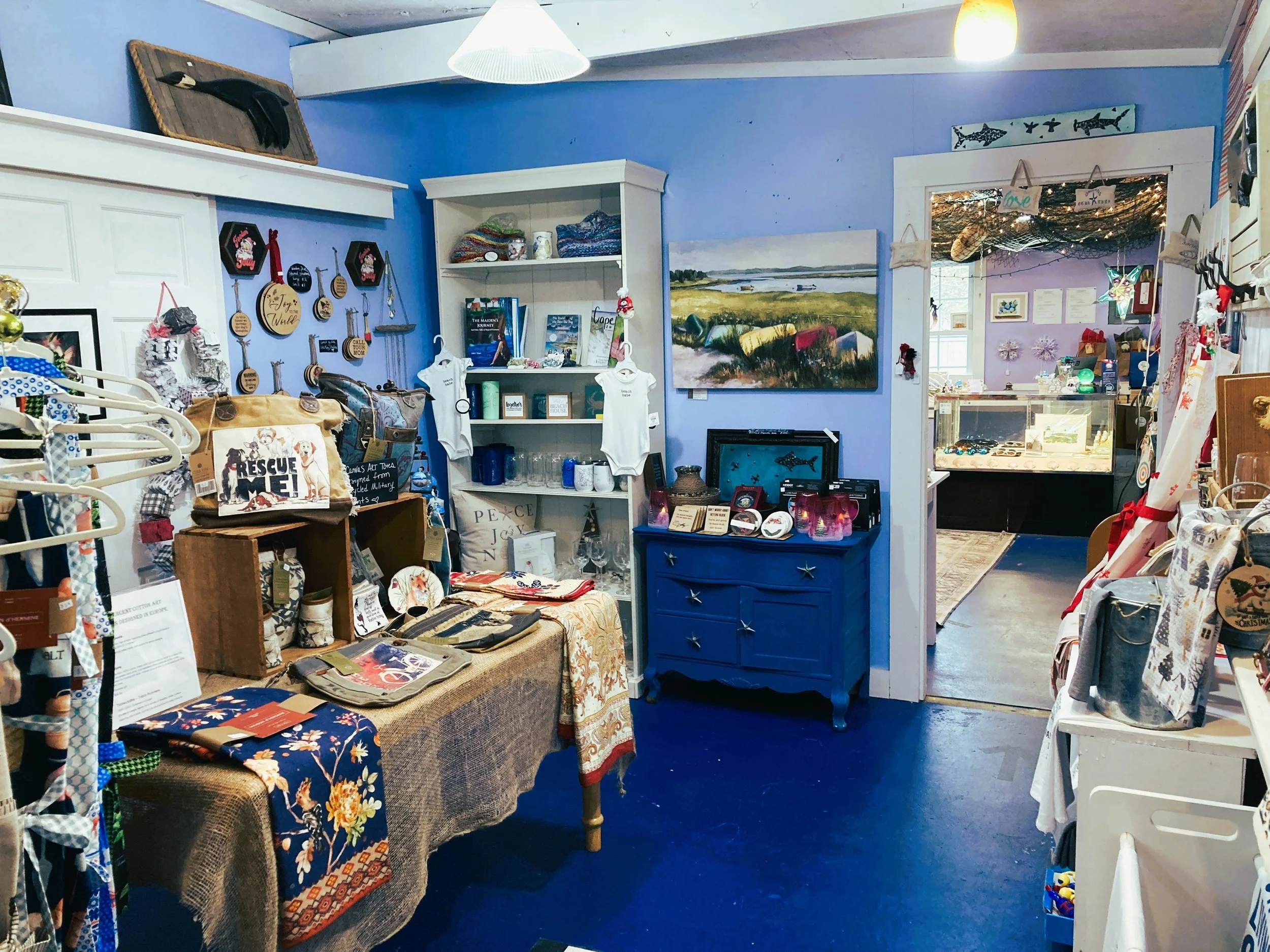 Inside a small boutique with various handmade and gift items, including paintings, jewelry, clothing, and decorative decorations, with vibrant blue walls and cozy lighting.