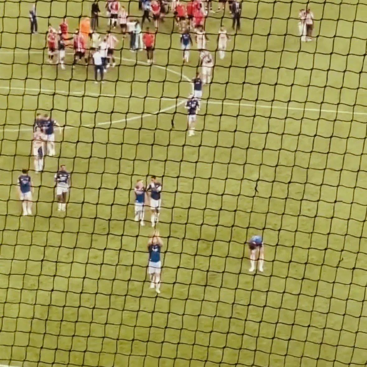 View of a soccer field with players and spectators, seen through a protective netting.