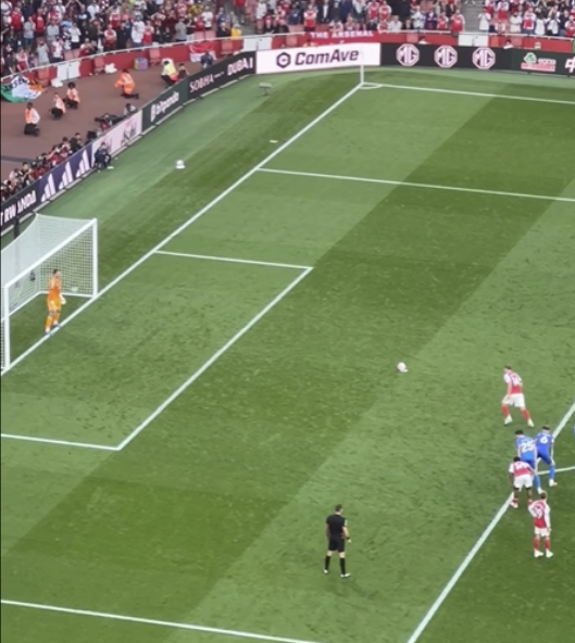 A soccer game scene showing a player preparing to take a corner kick, with the referee nearby, and four players near the goalpost. Spectators are visible in the stands.
