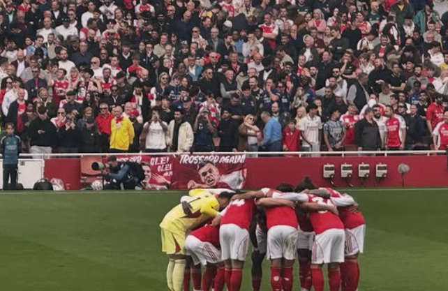 Soccer players huddled together on the field with an audience in the background.