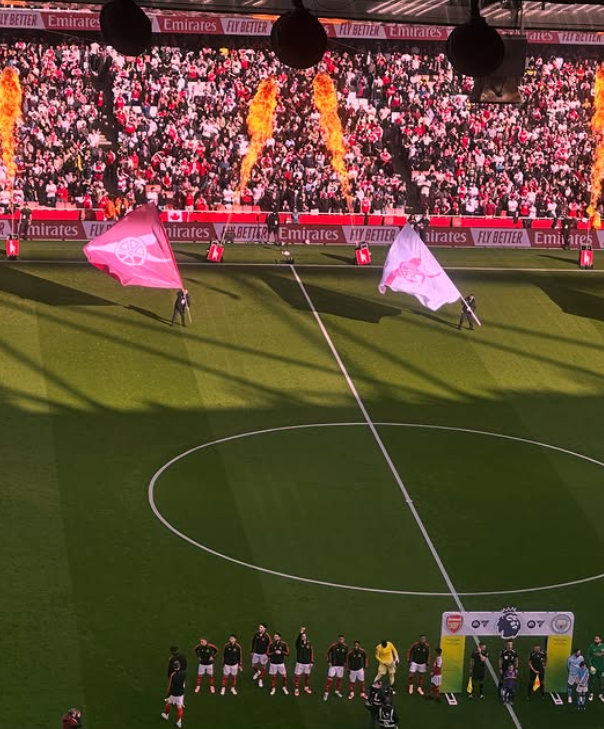Soccer stadium with players lined up at the field, flags being waved, and a large crowd in the stands. Fireworks are exploding in the sky.