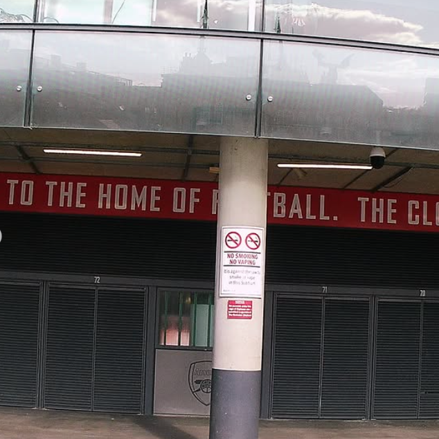 View of a stadium entrance with a sign that says 'No Smoking No Vaping' and a black and white column in front, with part of a red banner in the background.