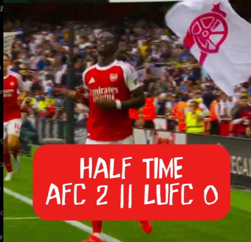Arsenal soccer player celebrating during match at halftime with fans and team staff in background, red scoreboard overlay showing halftime score of AFC 2, LUFC 0.