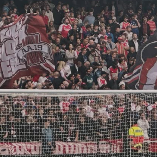 Crowd of football fans in the stadium stands, many wearing red and white, with large flags displaying team logos, and a security guard near the field.