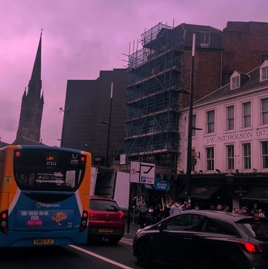 A city street scene with a bus, cars, and pedestrians. A building is under construction or renovation with scaffolding. A historic church steeple and a white historic building with 