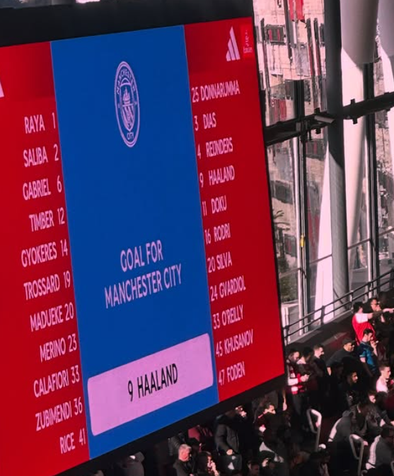 Large digital screen displaying Manchester City football team lineup for their match against Haaland, with a list of players on the left and right sides and team logo at the top center.
