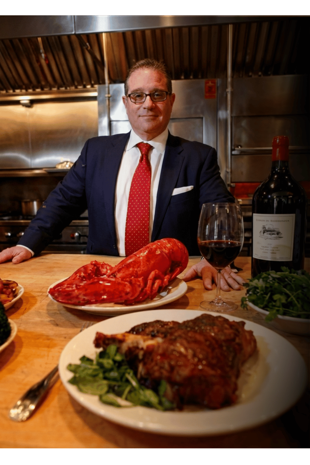 Chris Artinian in a suit and red tie standing behind a dinner table with various dishes, including a lobster, a glass of red wine, a bottle of wine, a roasted meat dish, and a salad, in a restaurant kitchen.