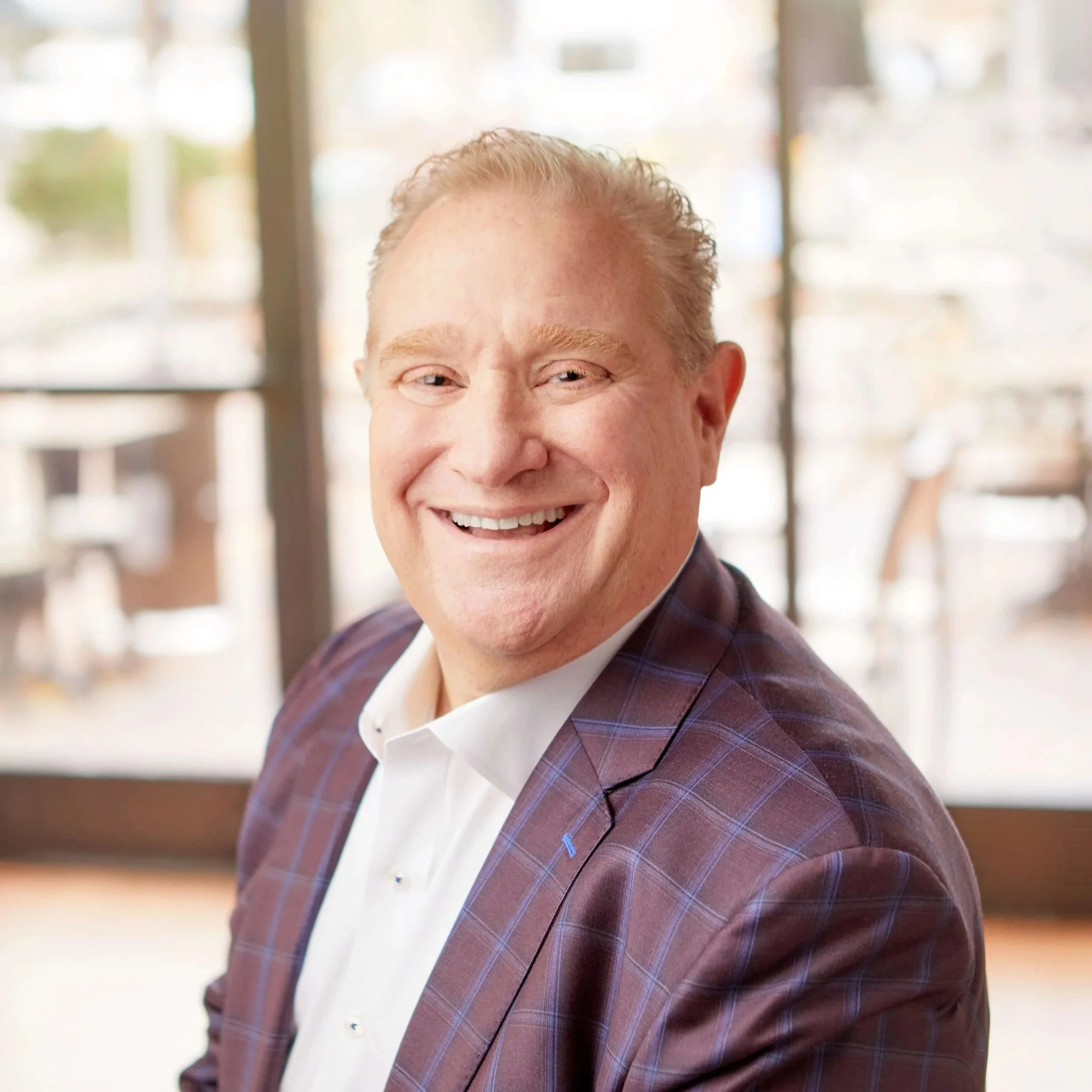 Chris Artinian wearing a white shirt and brown checkered blazer in an indoor setting with large windows in the background.