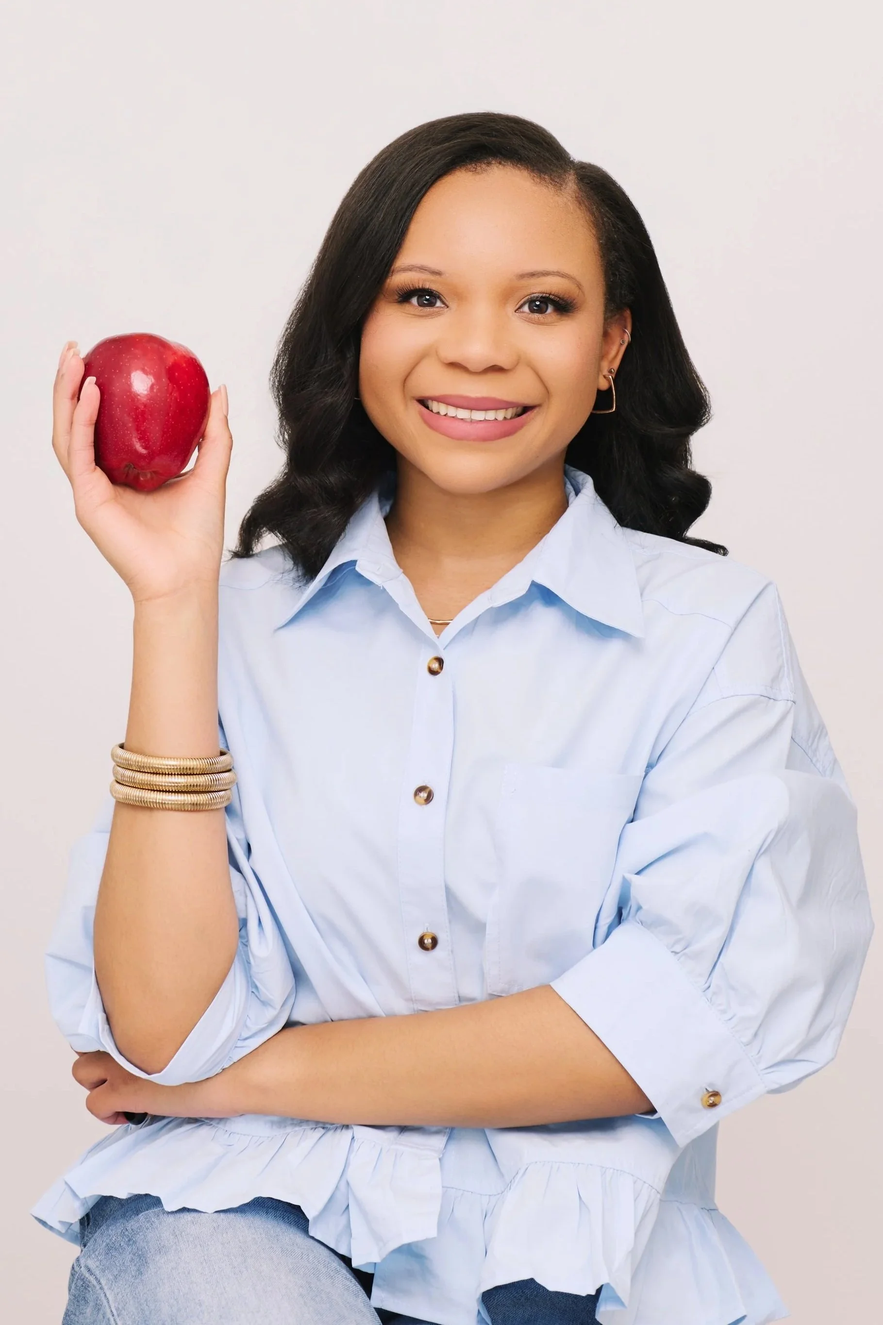 A young woman with dark, wavy hair styled in loose curls, wearing a light blue button-up shirt and gold jewelry, holding a red apple in her right hand and smiling at the camera against a plain white background.