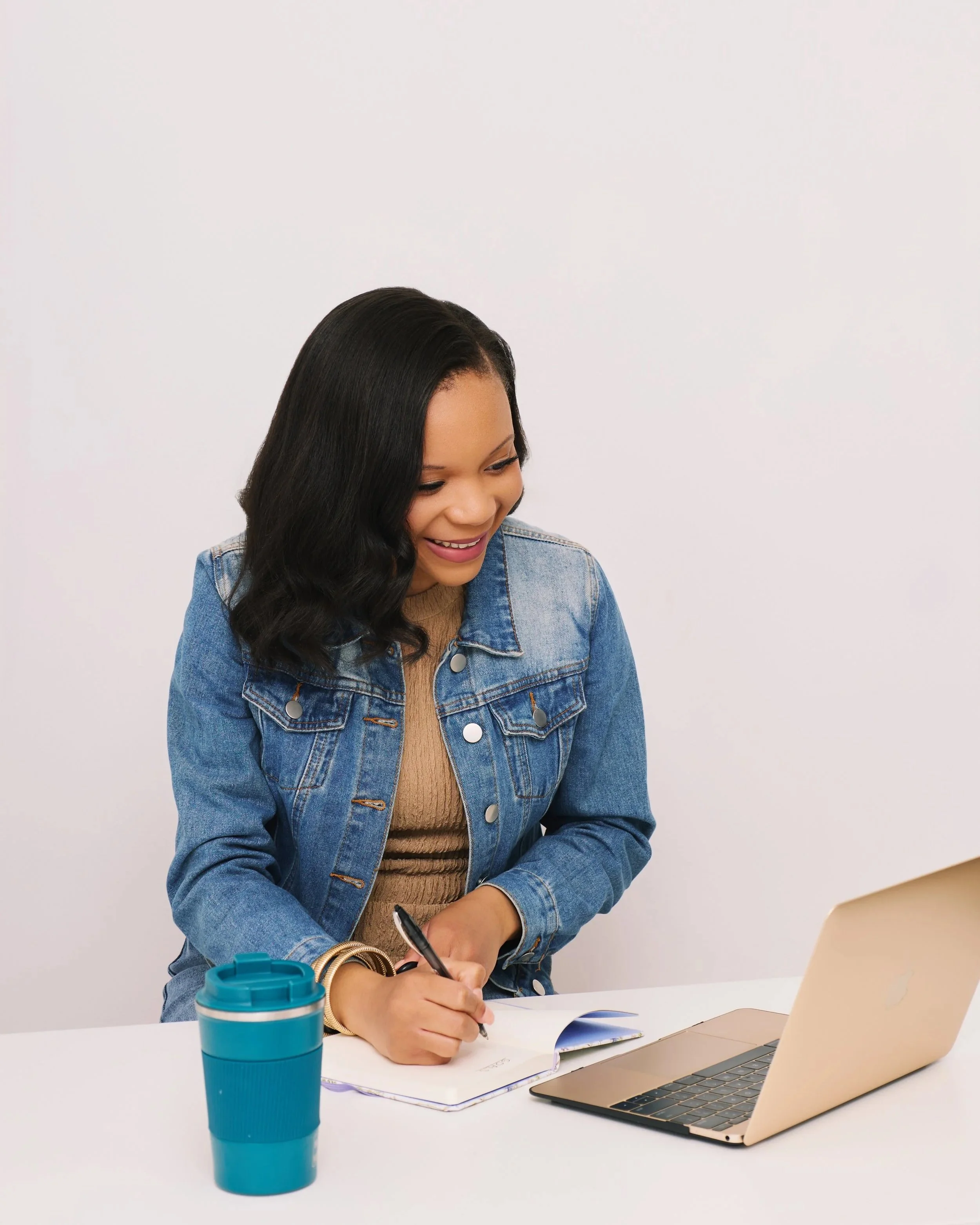 Young woman in a denim jacket writing in a notebook at a white table with a laptop and a blue travel mug.