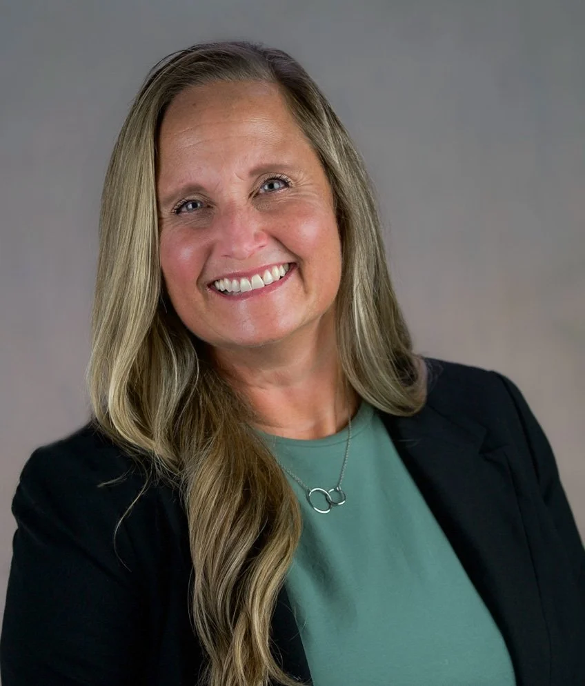 A woman with long blonde hair smiling, wearing a black blazer and a green top, with a silver circular necklace on a gray background.