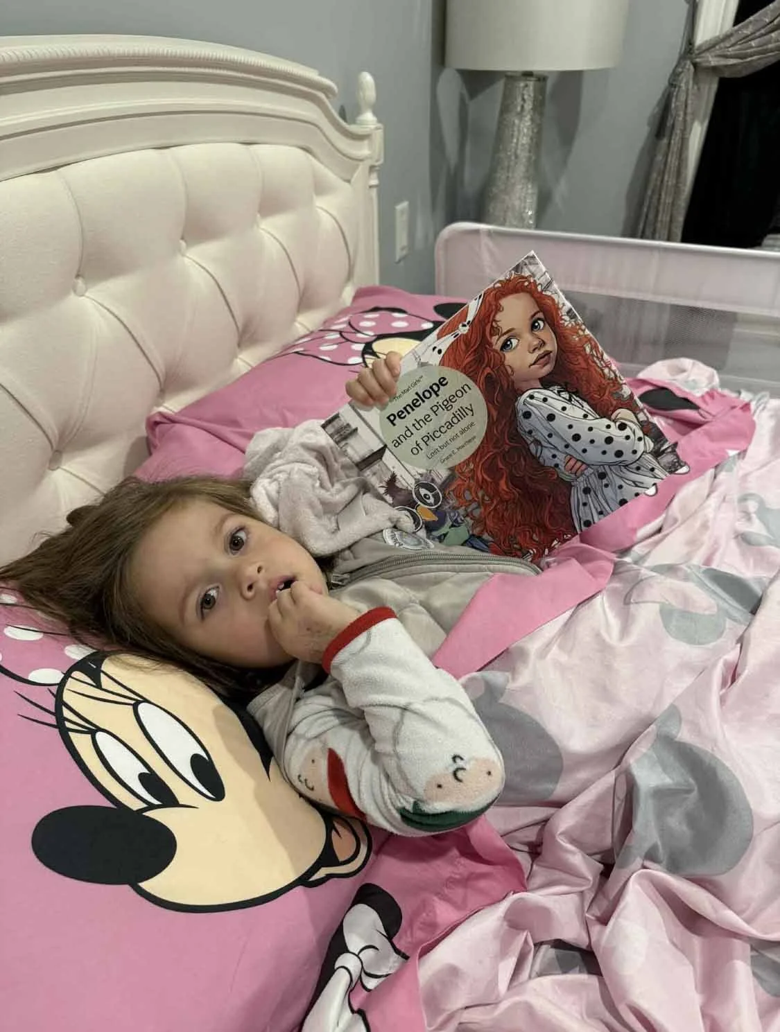 Two young girls lying in bed. One girl is resting her head on a Minnie Mouse pillow and the other is holding a book titled 'Penelope and the Pigeon of Piccadilly.' The bed has pink and gray Minnie Mouse themed bedding.