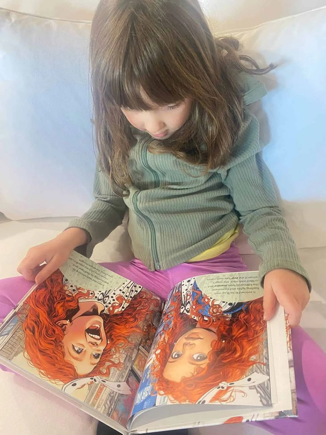 A young girl sitting on a bed, looking down at a comic book with illustrations of a woman with red hair.