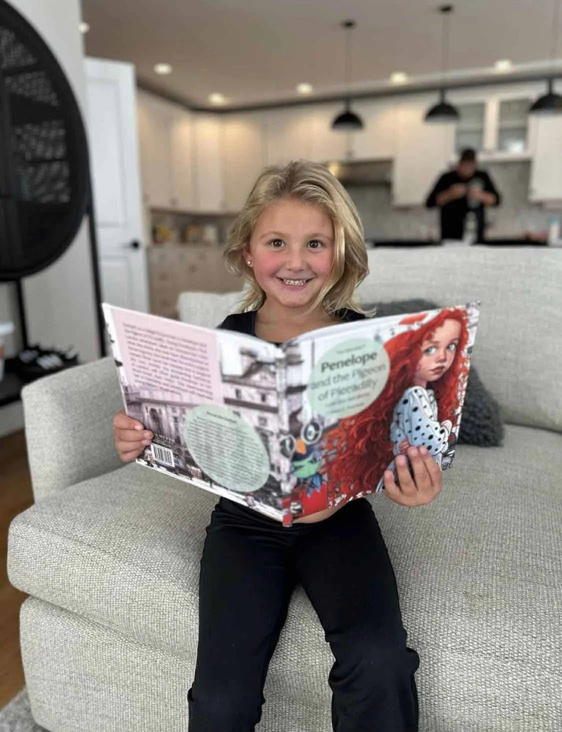 A young girl with blonde hair and a cheerful expression sitting on a beige sofa, holding a children's book titled 'Penelope and the Puzzle of Puzzl...