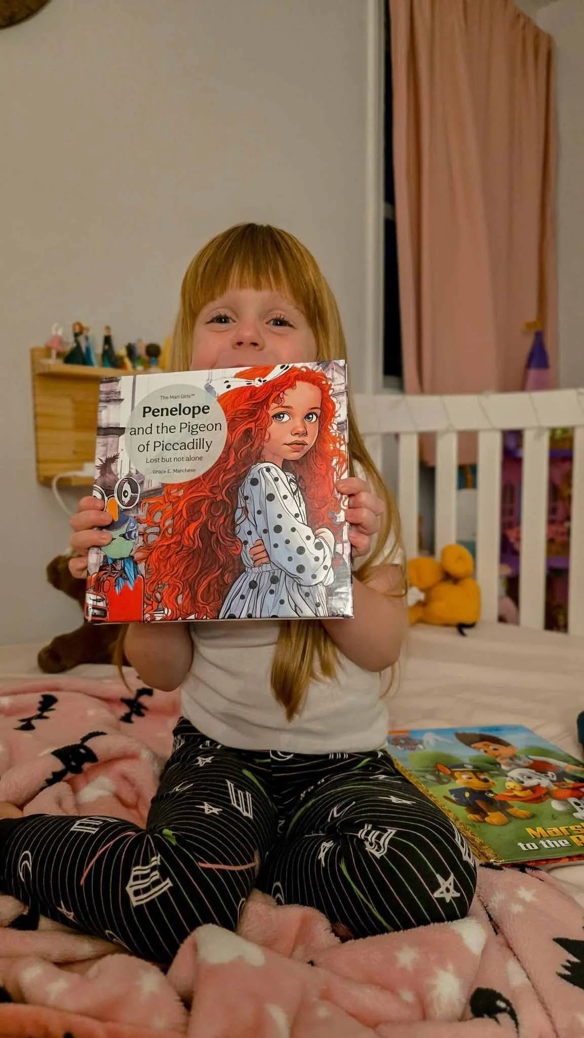 A young girl with red hair sitting on a bed, holding a children's book titled 'Penelope and the Pigeon of Piccadilly' in front of her face, with a pink blanket and a book about Paw Patrol nearby.