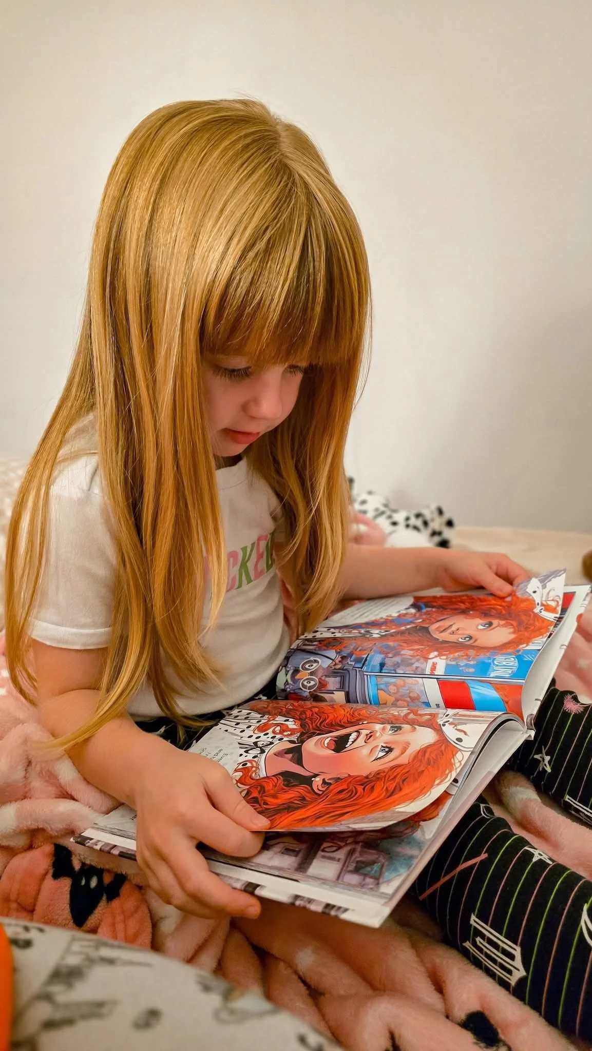 A young girl with long red hair reading a comic book while sitting on a pink blanket.