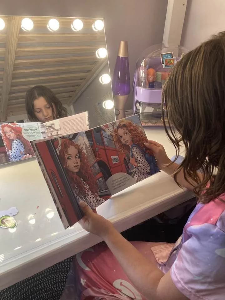 A young girl with glasses reading a colorful illustrated storybook at a vanity table. The mirror with lights shows her reflection, and there is a lava lamp and a container of items behind her.