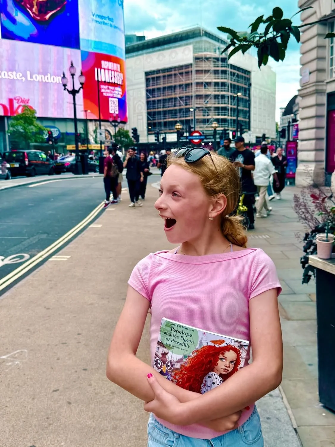 A young girl with red hair, wearing a pink t-shirt and holding a book, standing on a busy city sidewalk with advertising billboards, cars, and pedestrians in the background.