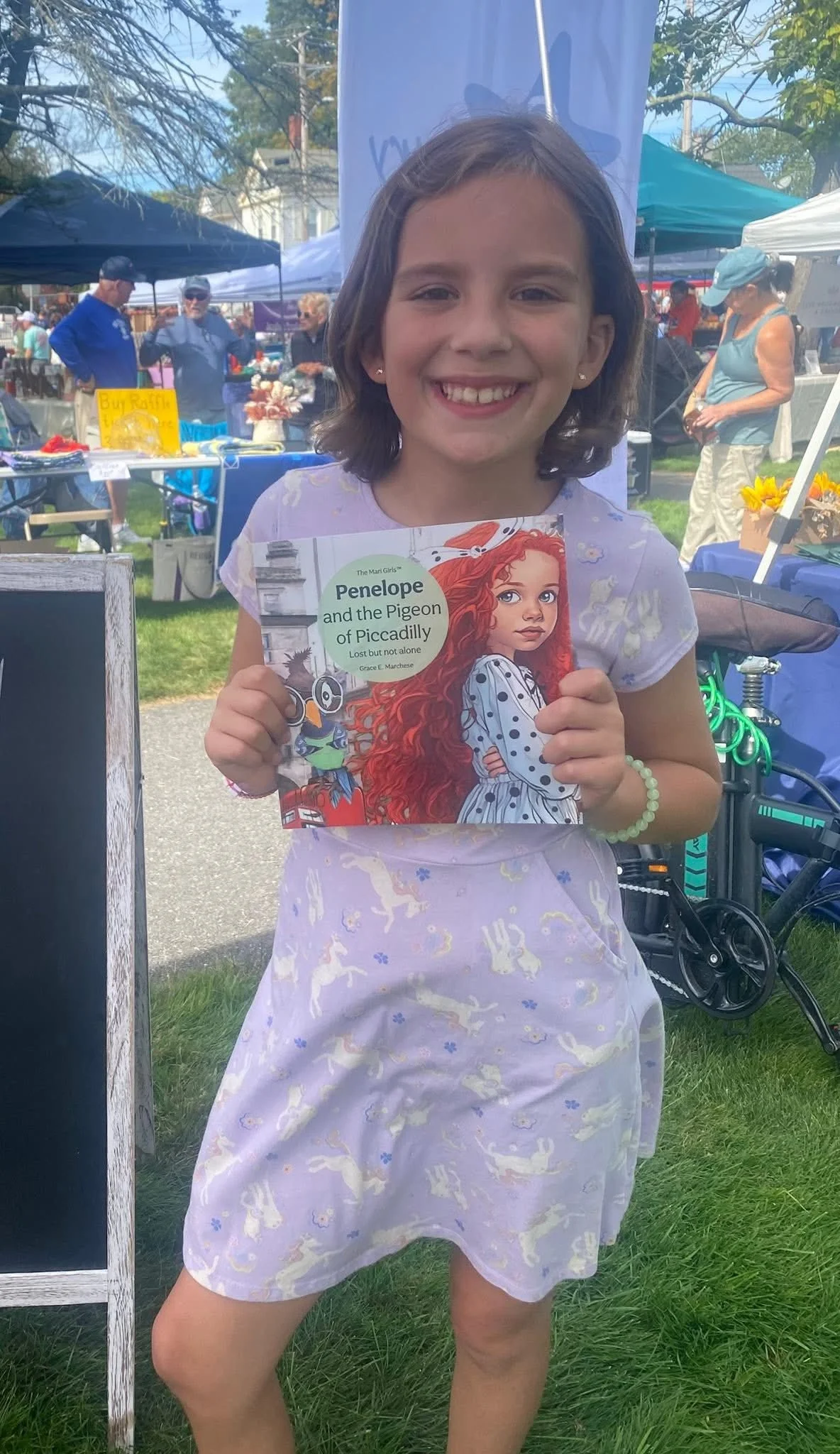 A young girl is smiling and holding a children's book titled "Penelope and the Pigeon of Piccadilly" at an outdoor market or fair, with tents and people in the background.