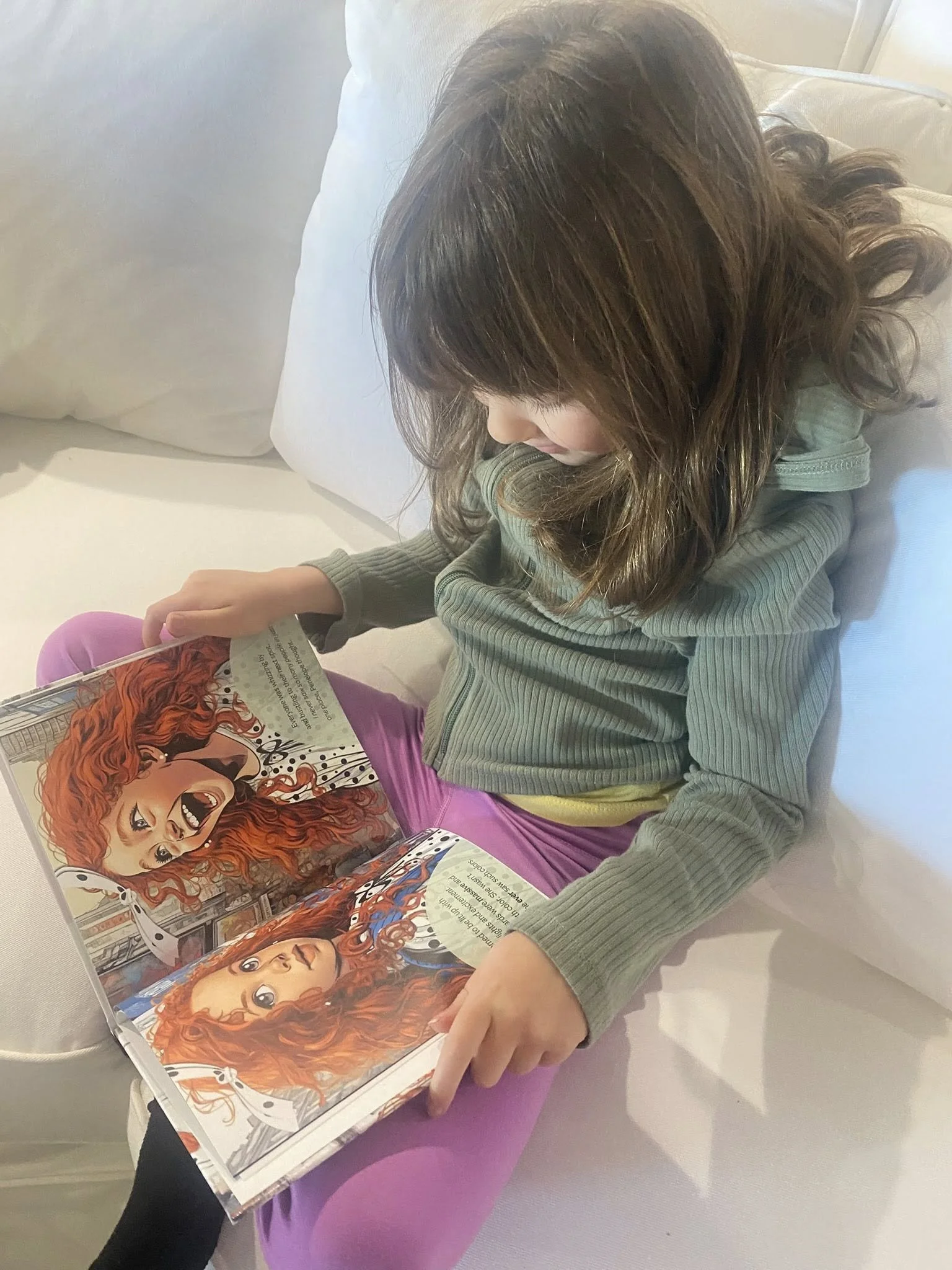 Young girl with wavy brown hair sitting on a white couch, reading a picture book with animated characters, including a girl with red curly hair.