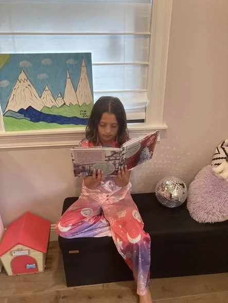 A young girl sitting on a black bench, wearing a colorful dress, reading a book. Behind her is a window with blinds, a painting of mountains and a lake, a silver disco ball, and a purple fuzzy cushion on the bench. A small red and white toy house is 
