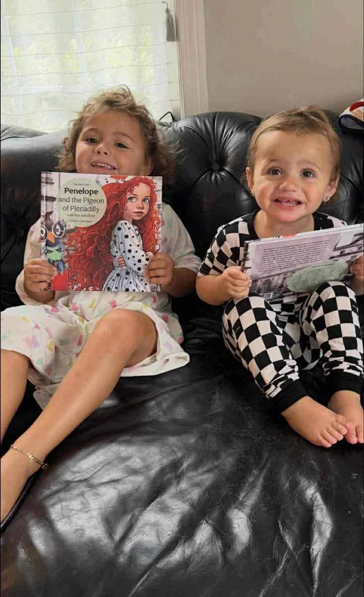 Two young girls sitting on a black leather couch, smiling and holding books. The girl on the left has curly hair, wearing pajamas with a floral pattern, and holding a book titled 'Penelope and the Pigeon of Piccadilly' featuring a red-haired girl on 