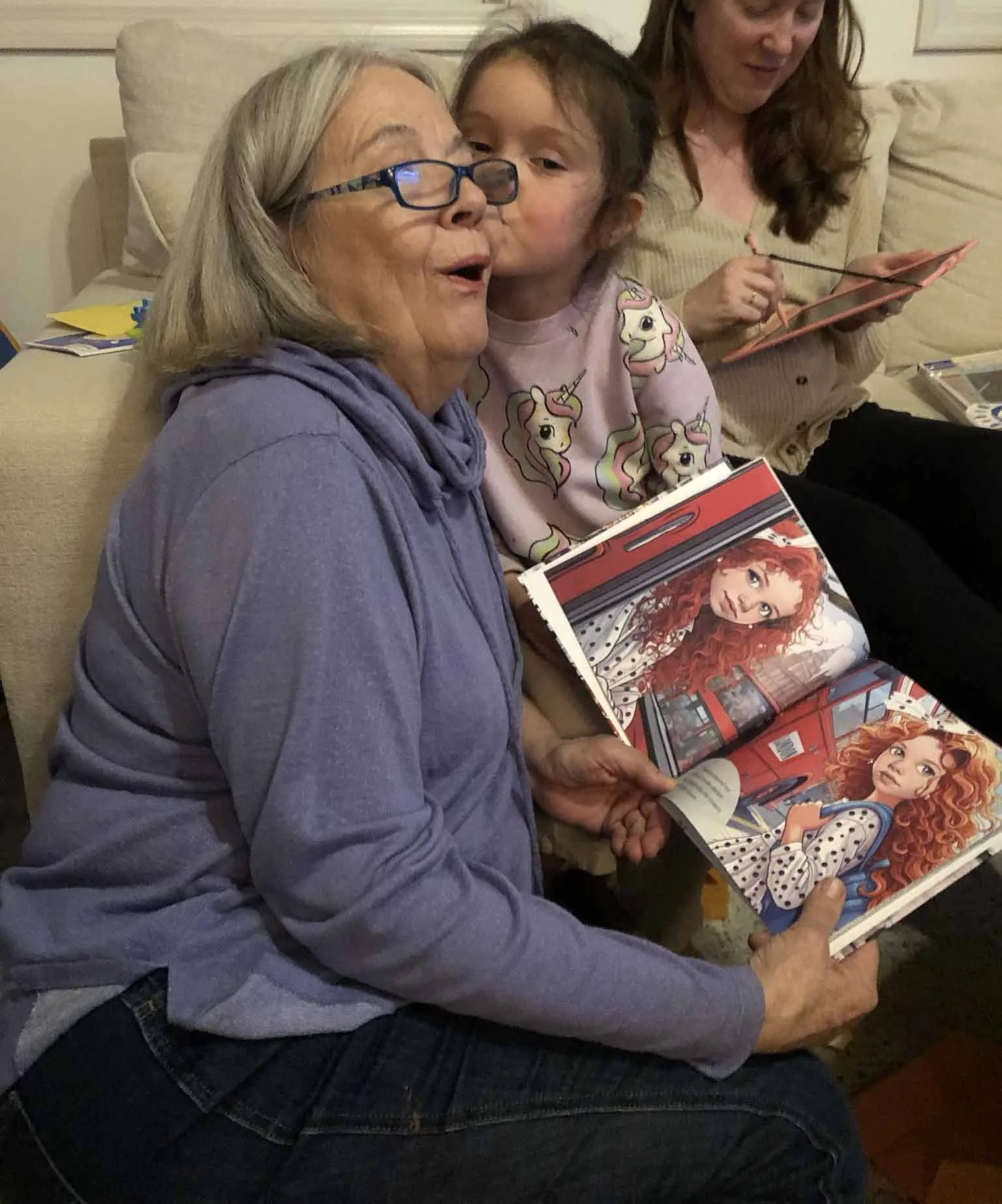 An elderly woman with gray hair and glasses sitting on a couch, holding an open comic book or magazine featuring a red-haired girl with green eyes and a polka-dot dress. A young girl is giving her a kiss on the cheek, and a woman in the background is