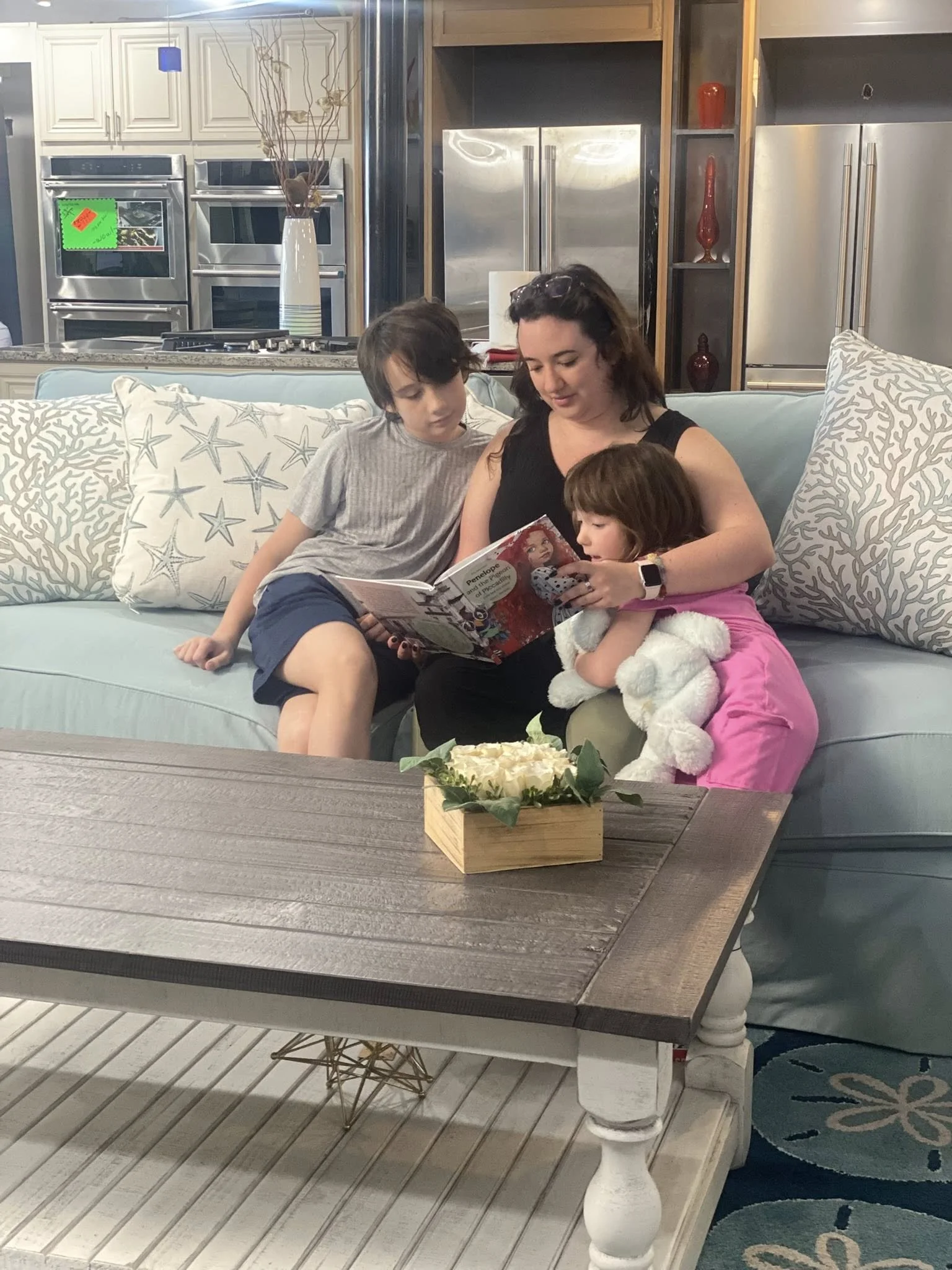 A woman with two children sitting on a light blue sofa in a living room, looking at a magazine. One child is a girl holding a plush toy, and the other is a boy leaning in to look. The coffee table in front has a small decorative plant. The background