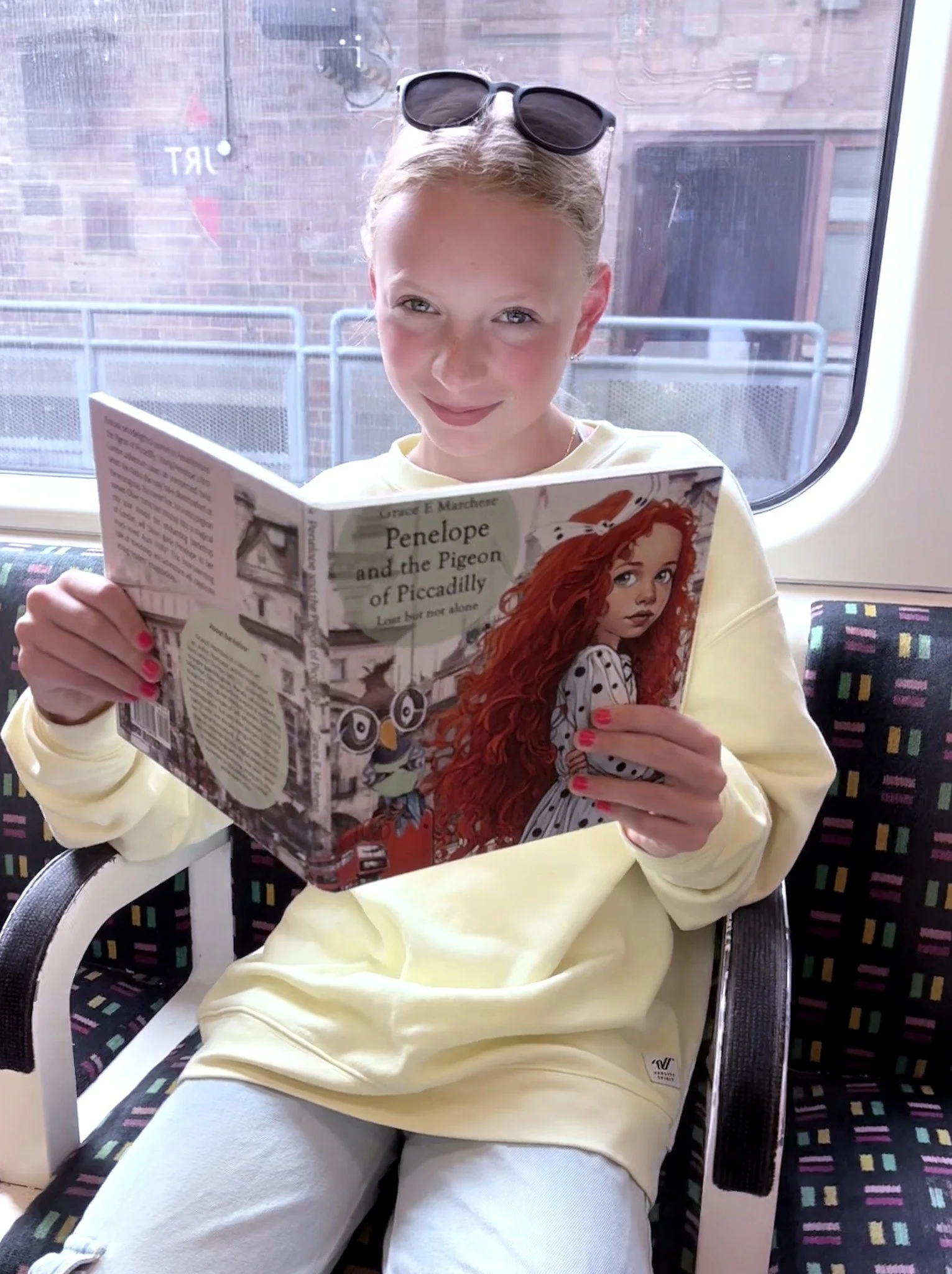 A young girl with blonde hair and sunglasses on her head is sitting on a train seat, reading a book titled 'Penelope and the Pigeon of Piccadilly'. She is wearing a light yellow sweatshirt and light-colored pants, with pink nail polish.