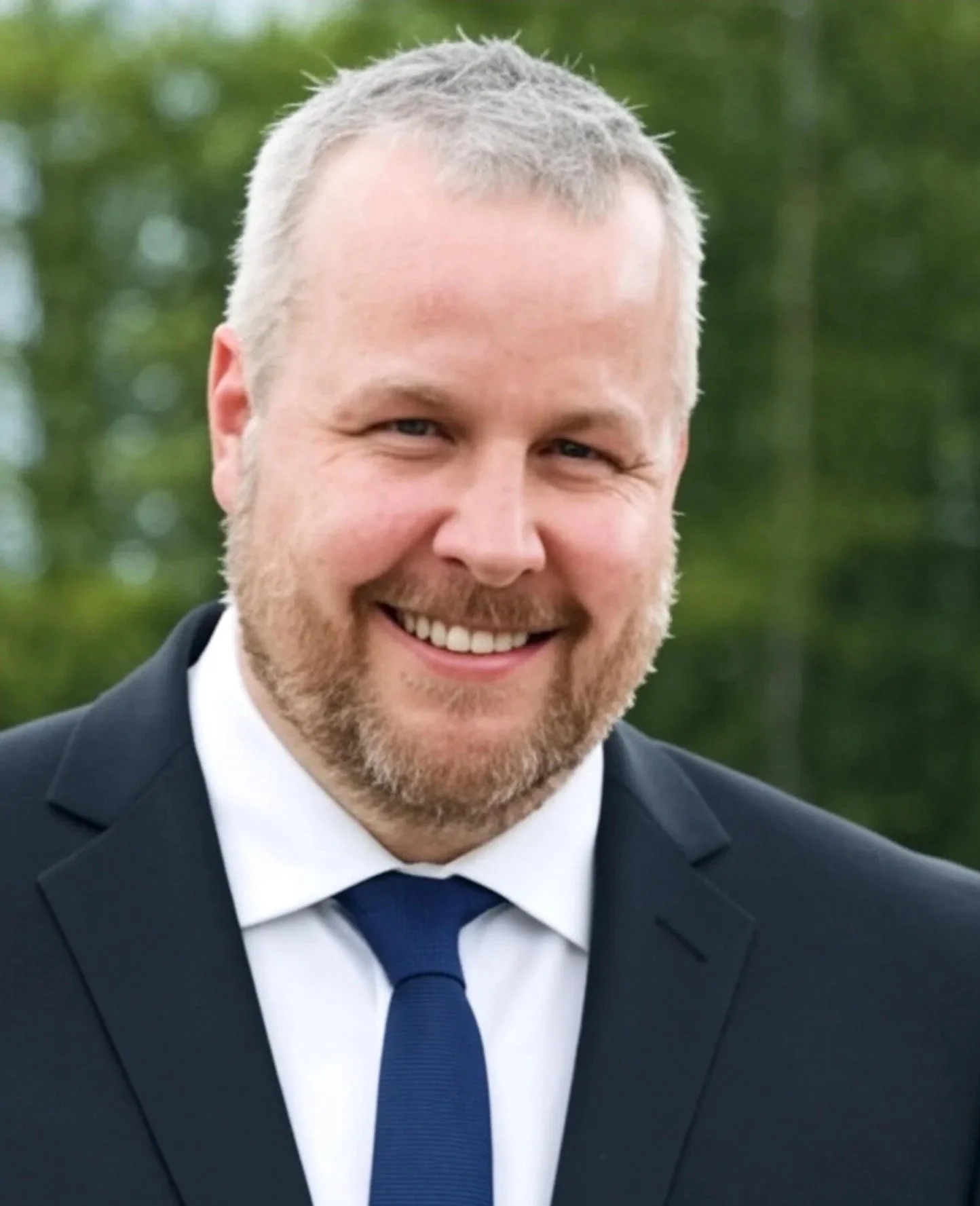A man with short, light-colored hair and a beard wearing a dark suit, white shirt, and dark blue tie, smiling outdoors with a blurred green background.