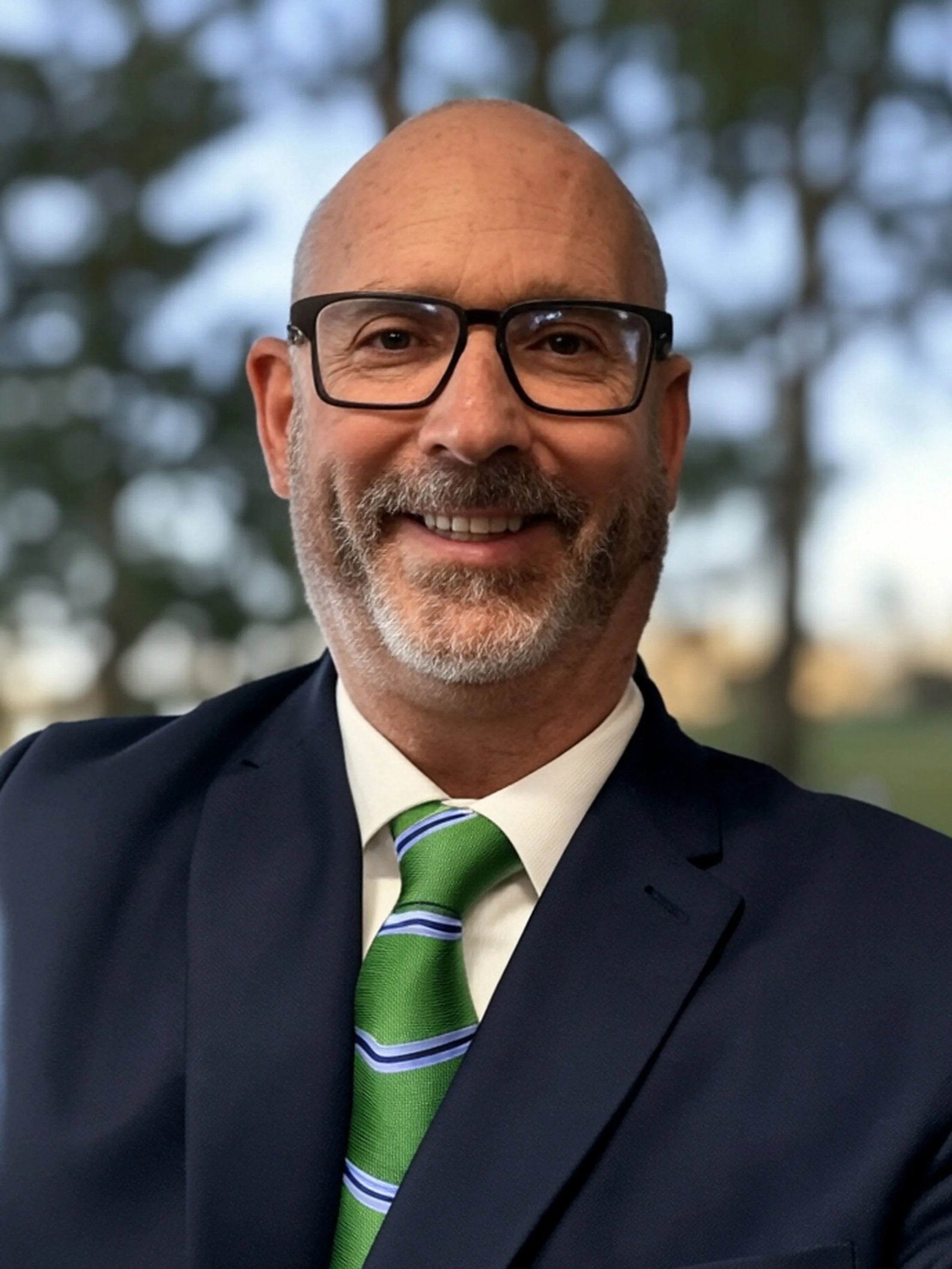 A smiling bald man wearing glasses, a navy suit, a white shirt, and a green striped tie, standing indoors with a blurred background of windows and hanging green items.
