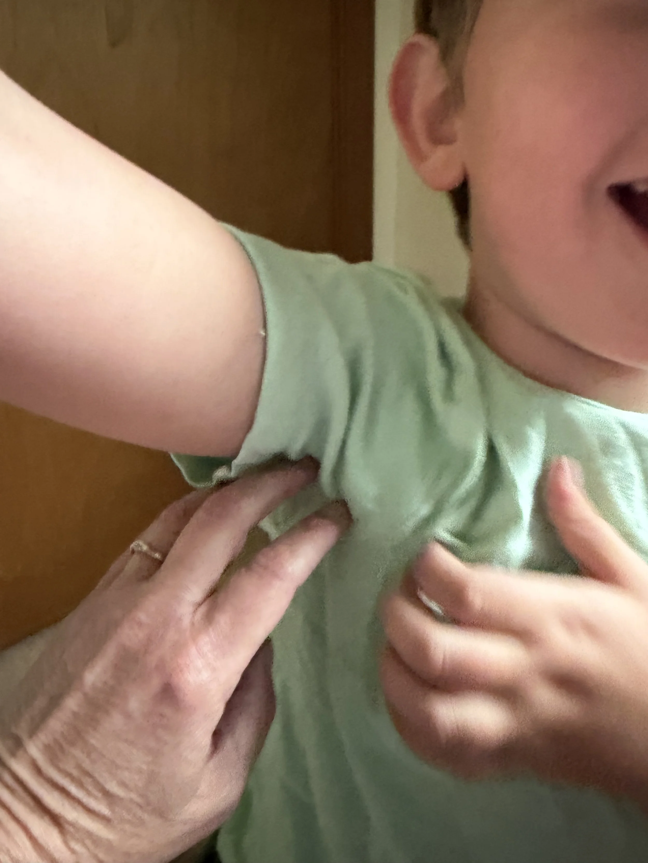 The upper body of a five-year-old child with pale skin wearing a light green shirt is shown, raising an arm while one adult hand and one child's hand tickle the exposed armpit.