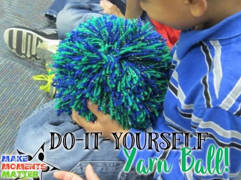 Child holding a yarn ball made from multicolored yarn with a blue, green, and glow-in-the-dark pattern, sitting on the floor with other children nearby, with text promoting a DIY yarn ball activity.