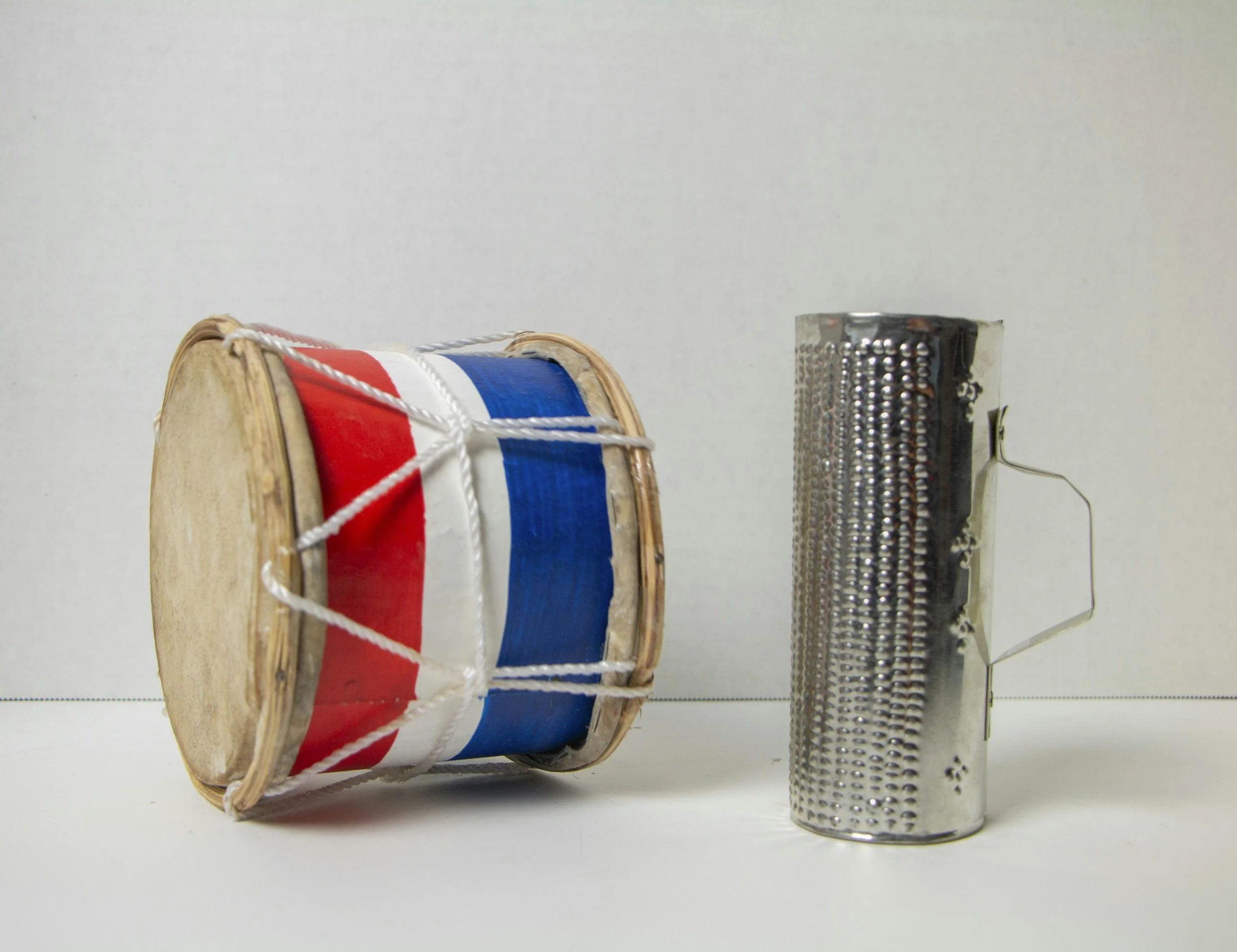 A small red, white and blue hand drum and a metal guiro standing vertically on a white surface.