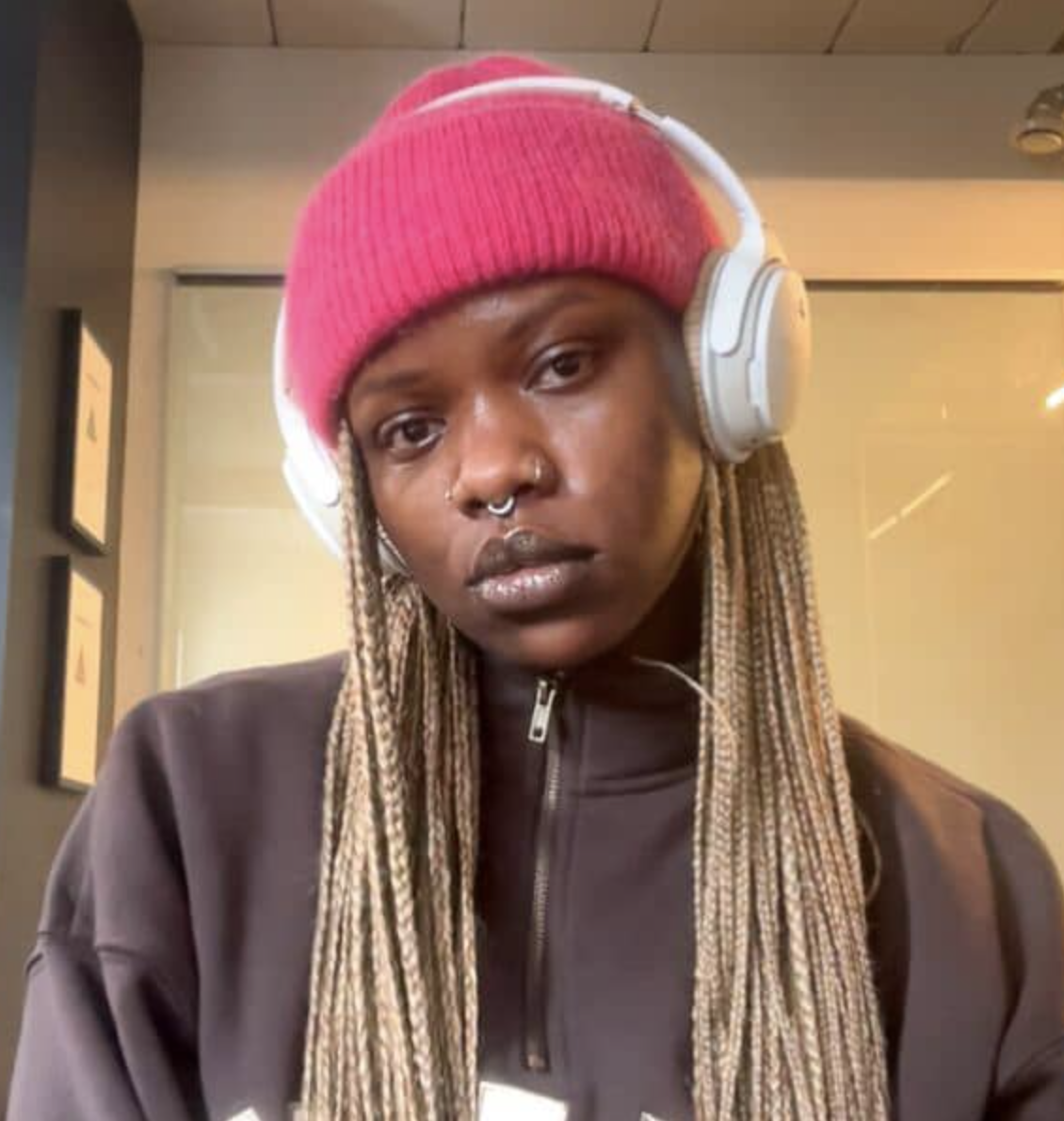 A young woman with brown skin wearing a pink beanie and white over-ear headphones, with a nose ring and dreadlocks, looking directly at the camera in an indoor setting.