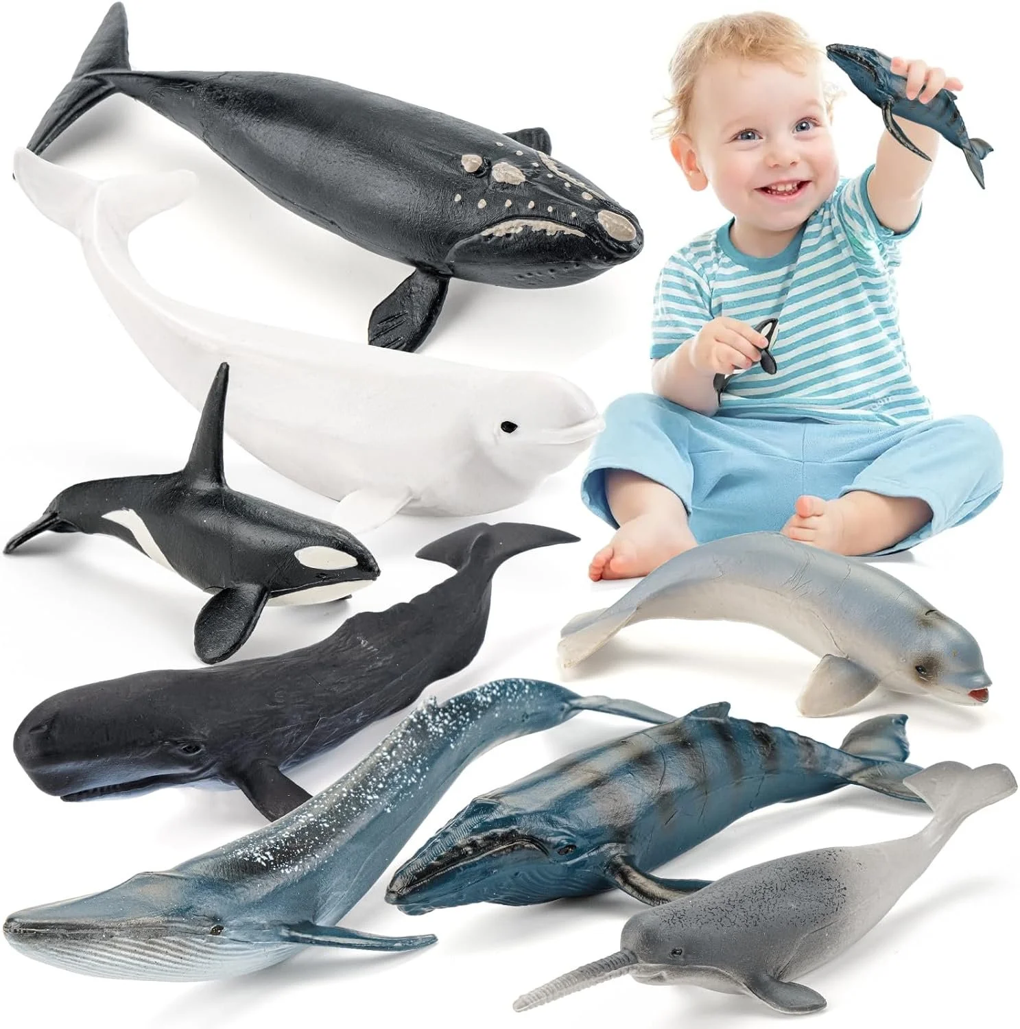 A young boy sitting on the floor surrounded by toy sea animals, including whales, sharks, and dolphins, holding a small fish toy and smiling.