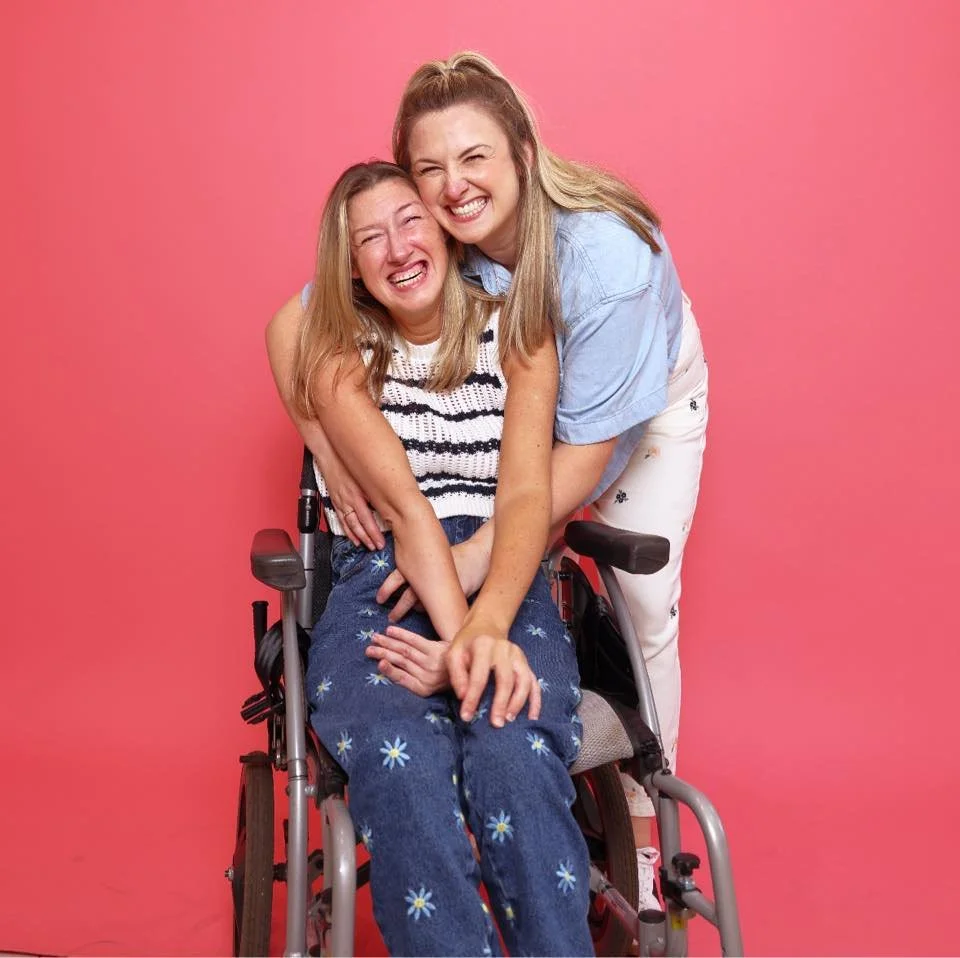 Two women laughing and hugging, one sitting in a wheelchair, against a pink background.