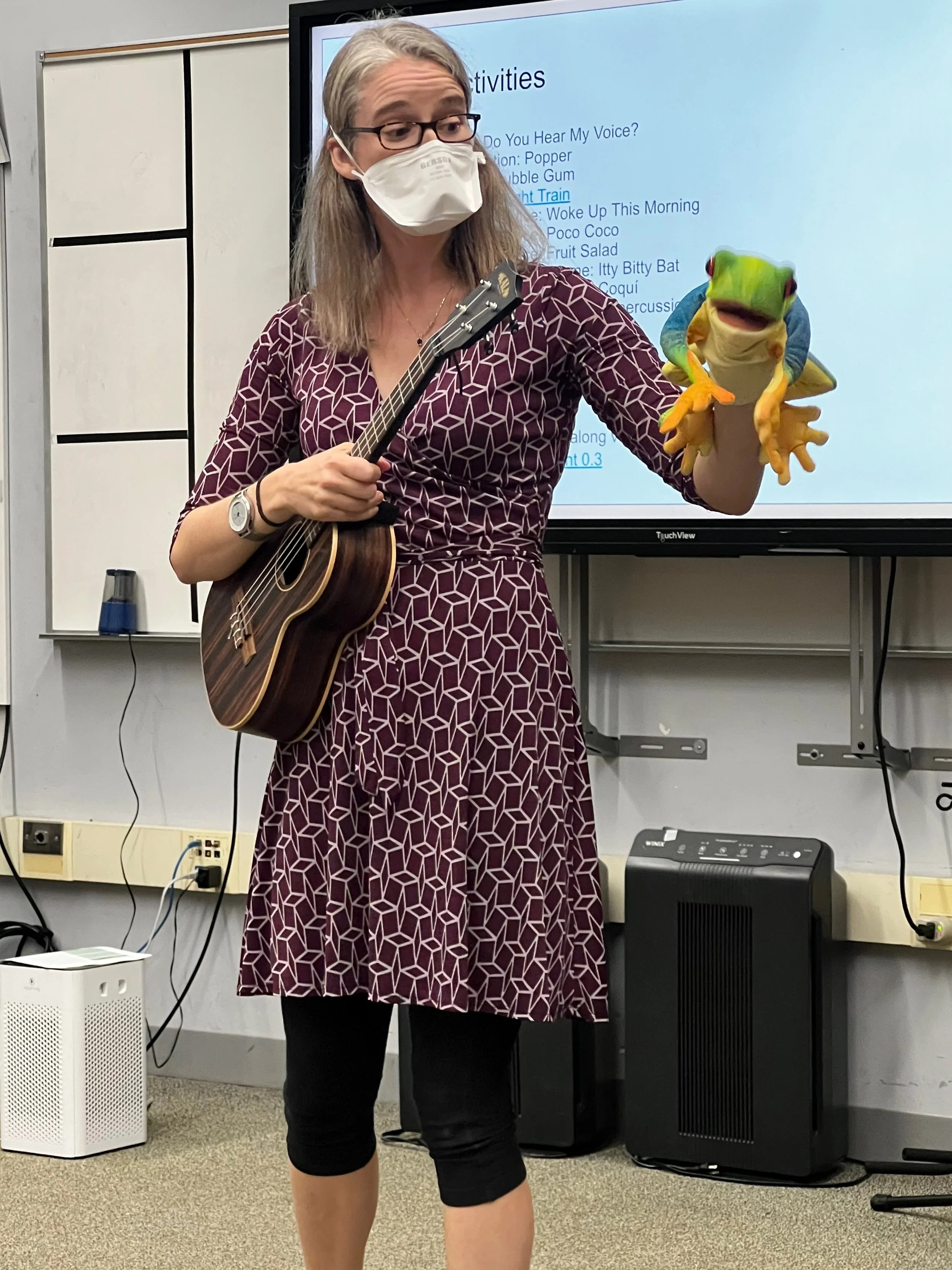 Jessica Corwin presents to a teacher workshop. She is wearing a patterned purple dress, a duck bill style face mask, a ukulele and a frog puppet on her left hand.