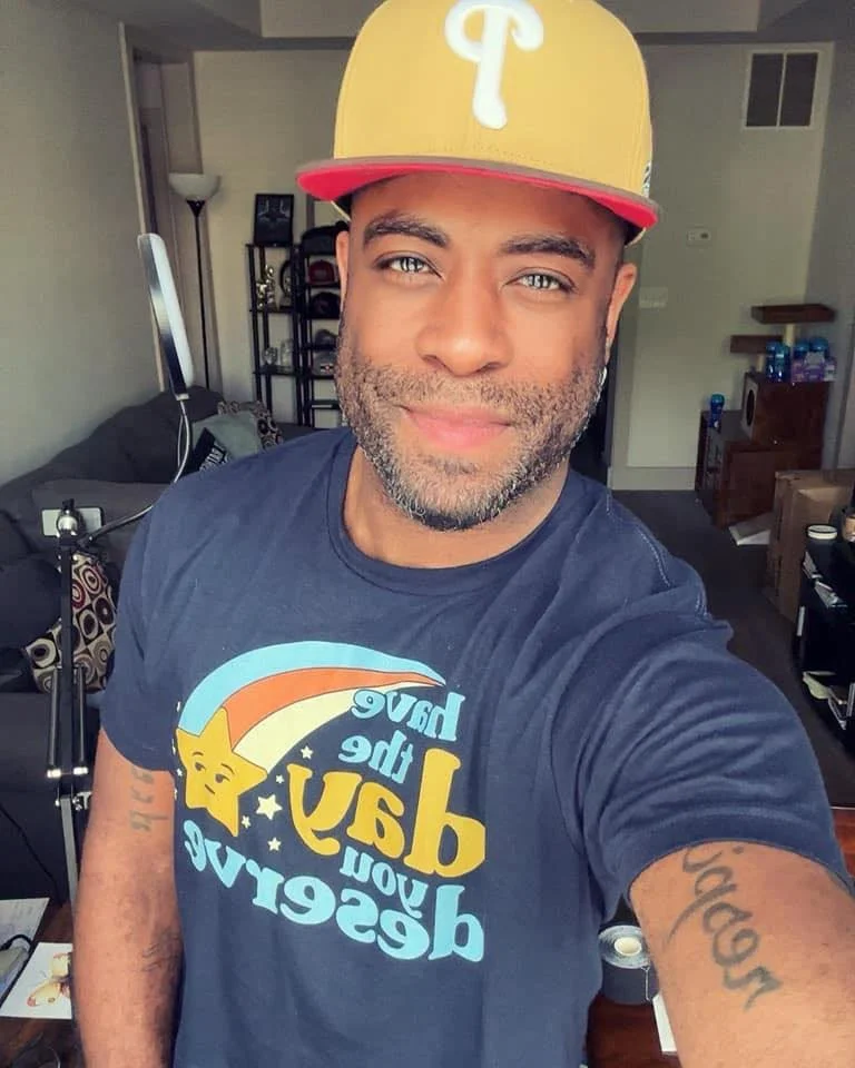 A man smiling for a selfie inside a living room, wearing a beige cap with a white emblem and a dark T-shirt with a colorful design and text that reads 'Have the day you deserve'.