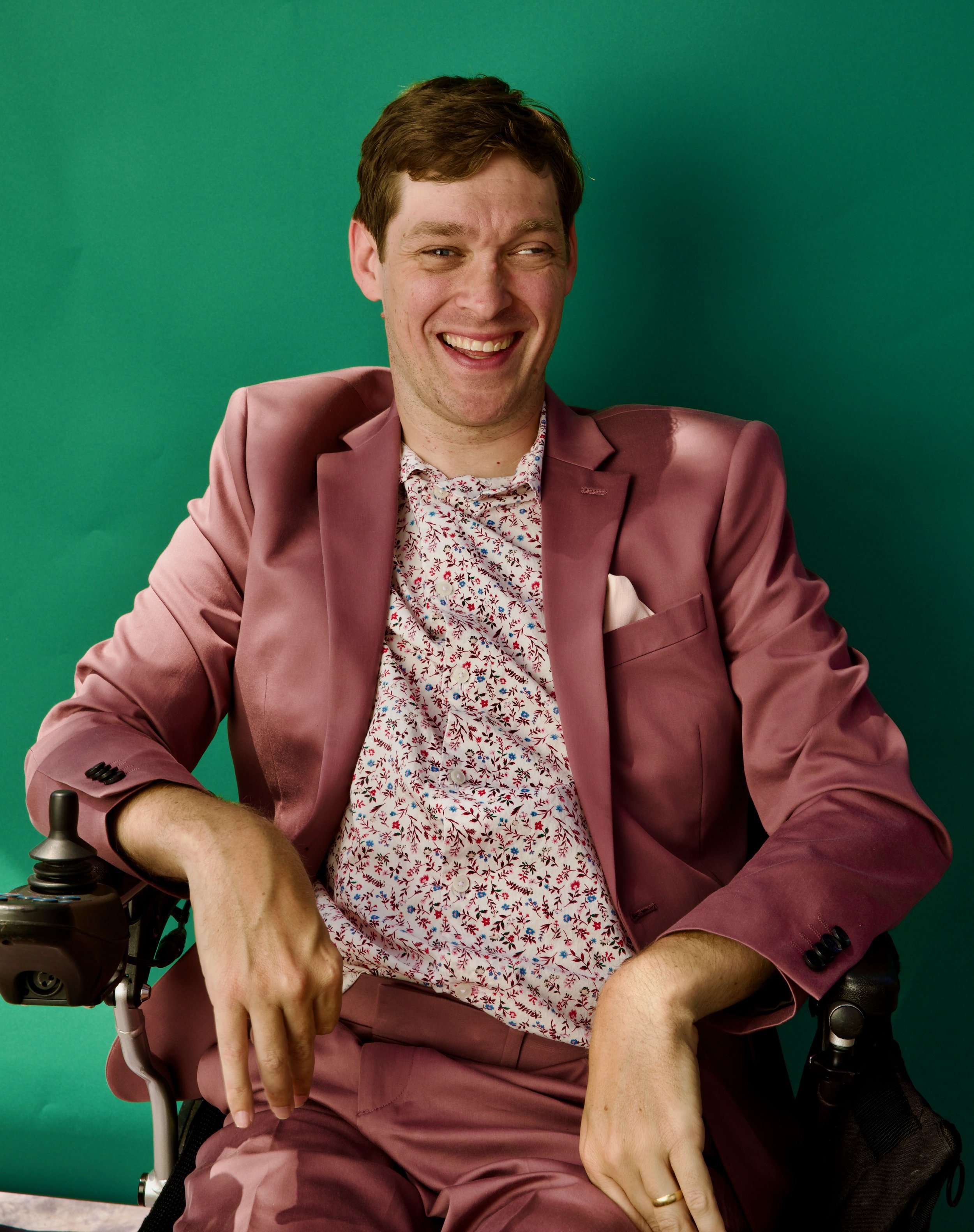 A smiling man in a pink suit and floral shirt sitting in a wheelchair against a green background.
