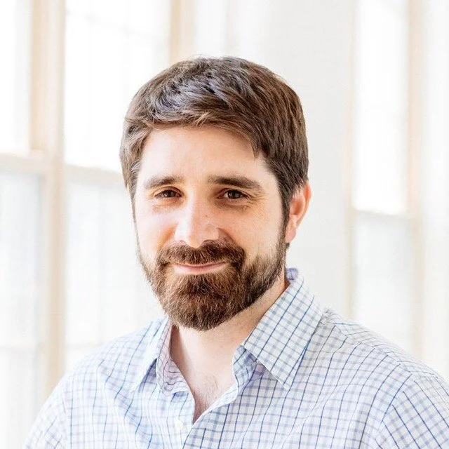 Philip Silva is standing in front of a wall of white windows and gently smiling at the camera. He has dark brown hair and beard and is wearing a white shirt with blue grid lines.