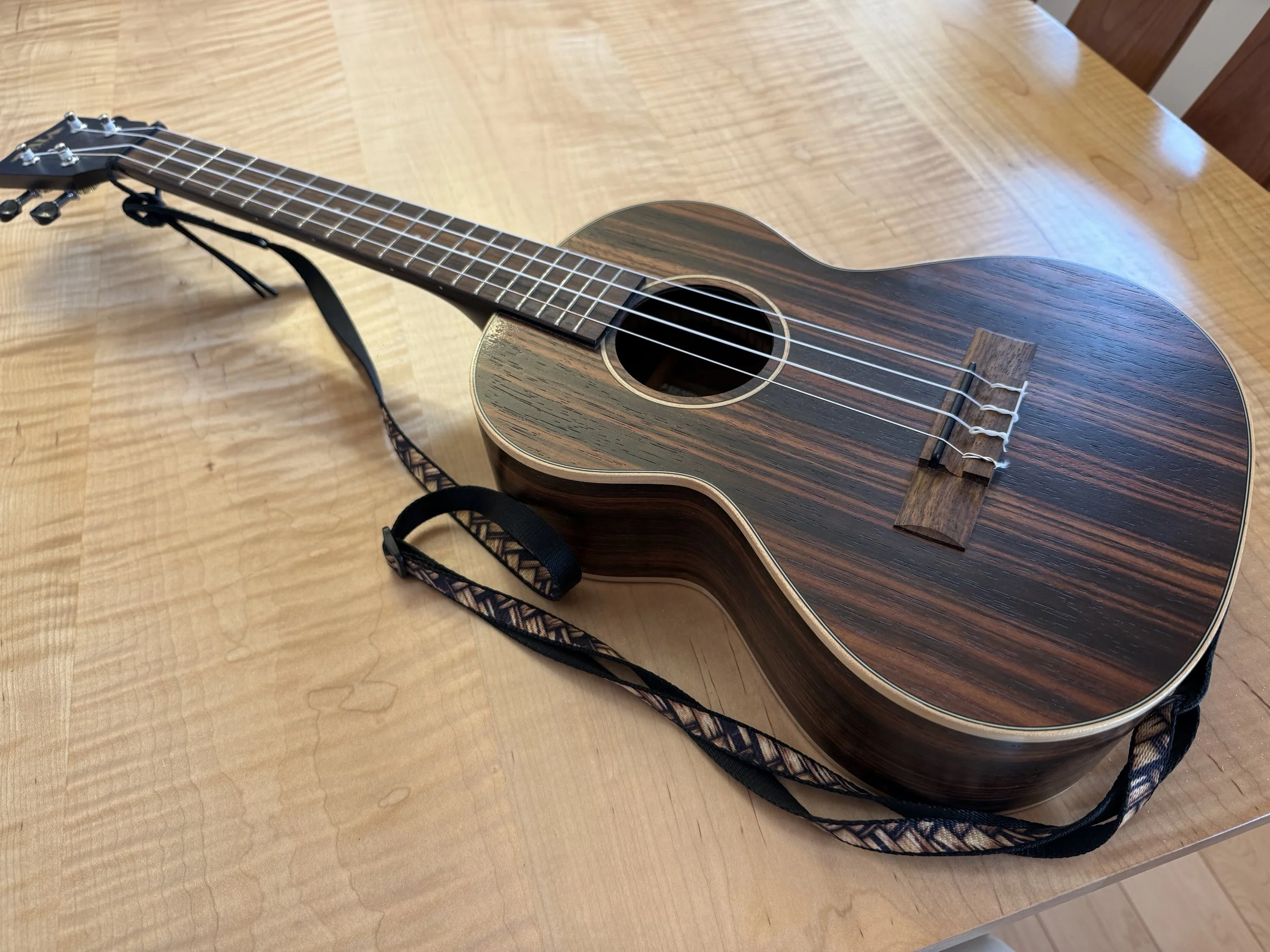 A dark brown wooden ukulele with light colored edges and a skinny black and brown shoulder strap sits on a light maple tabletop.