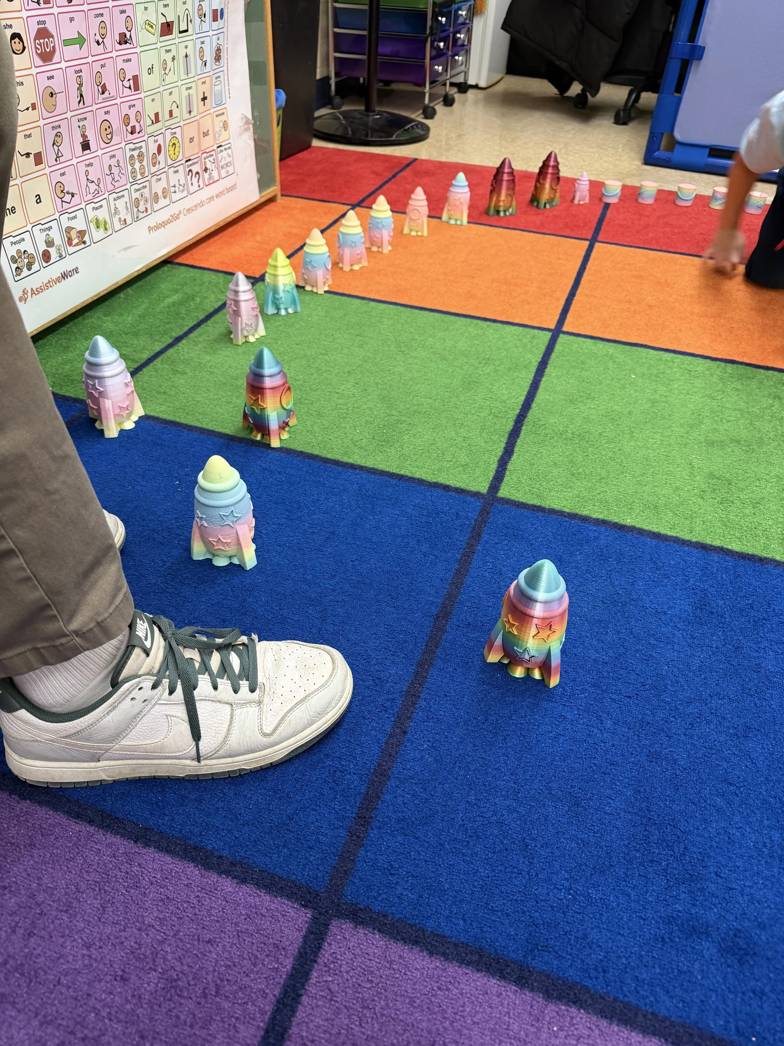 Colorful rocket shakers arranged in a grid on a multicolored carpet in a classroom.