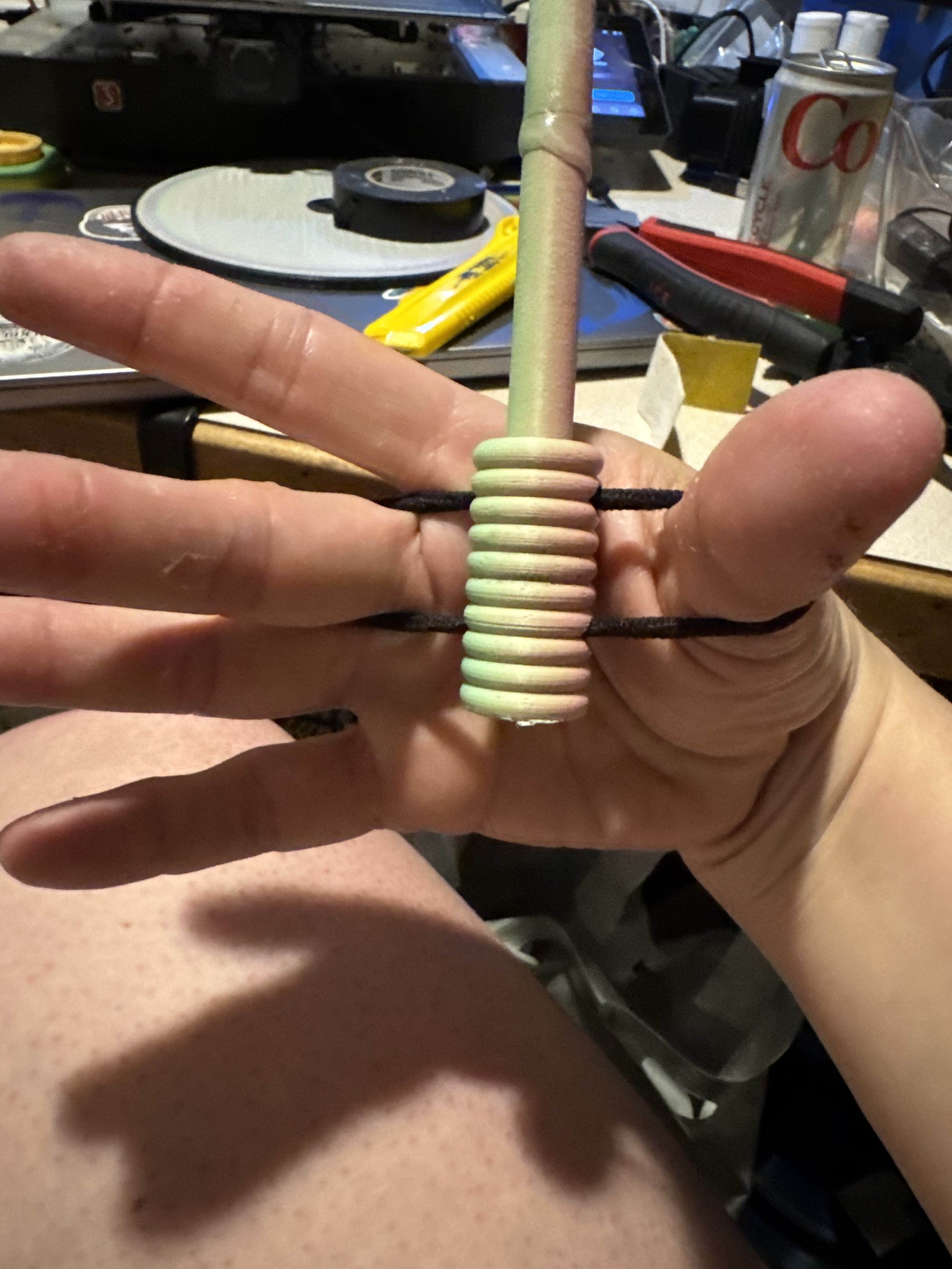 A percussion striker with attached elastic is modeled in an open hand that does not need to grasp the stick tightly to operate the instrument. The background is cluttered.