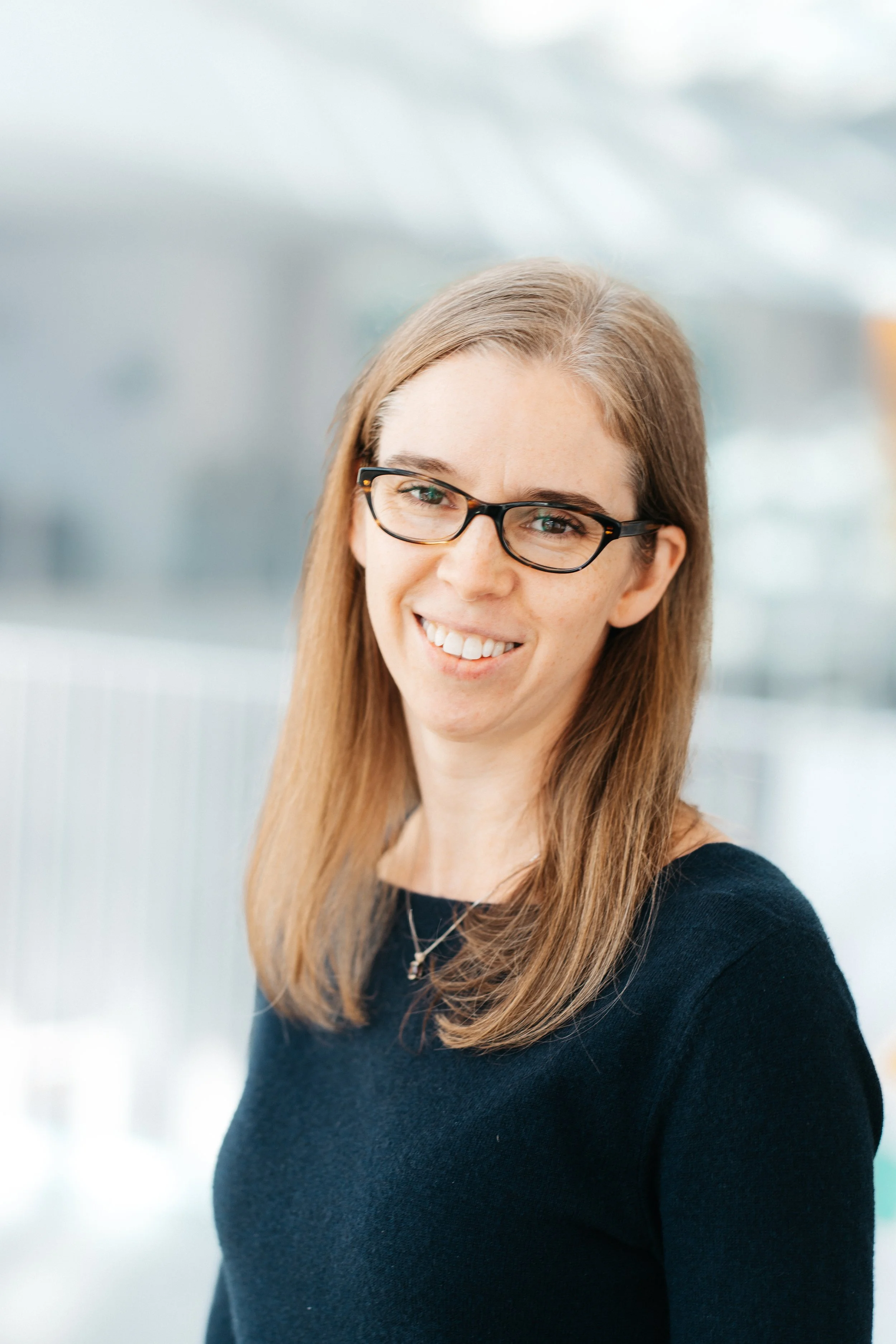 Jessica Corwin is a white woman with brown glasses and straight light brown hair past her shoulders, smiling at the camera. She is wearing a navy blue dress.