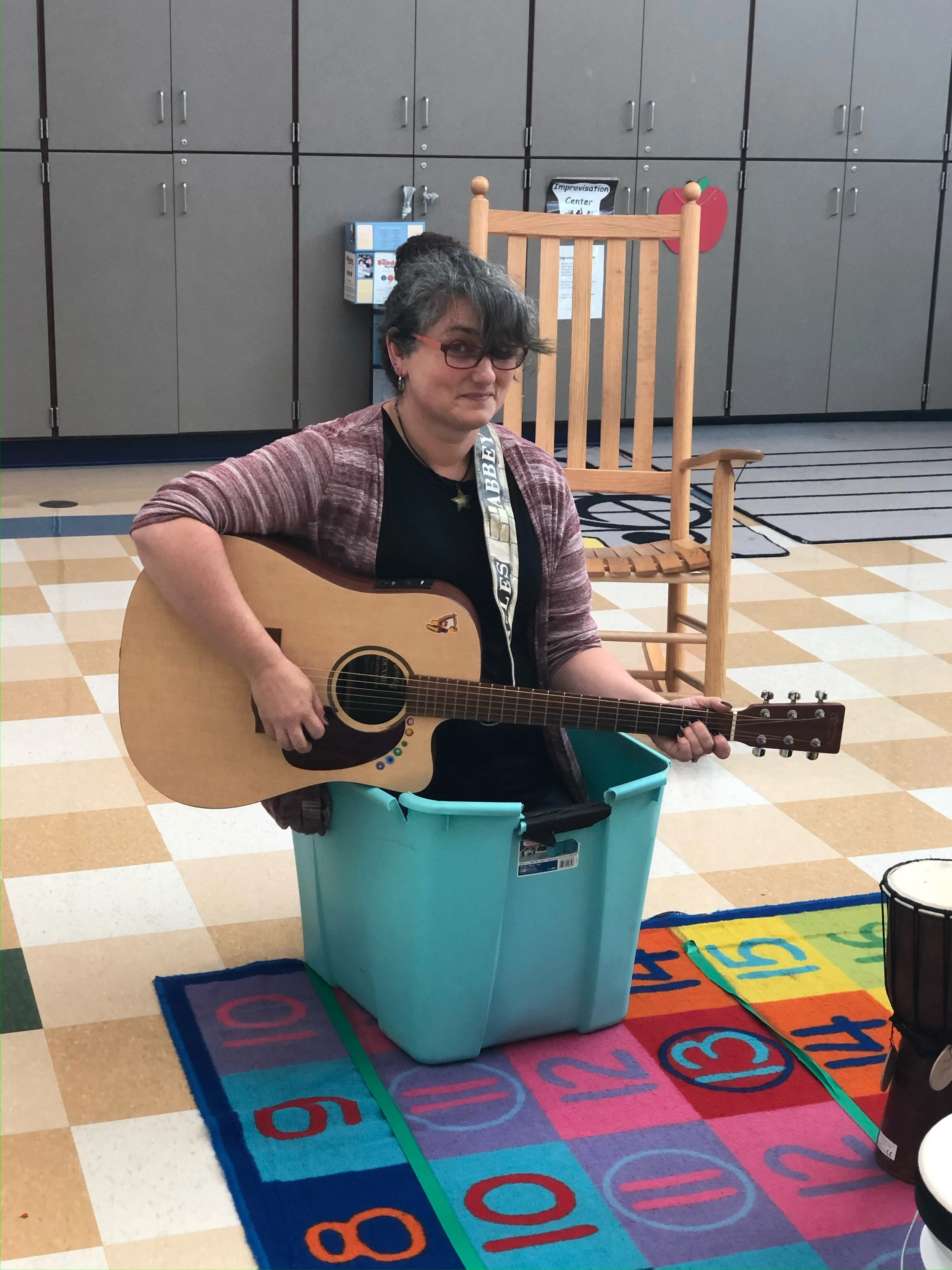 Jen Kass sits in a turquoise storage tub on the floor of a classroom playing guitar. She is wearing a striped sweater and smiling.