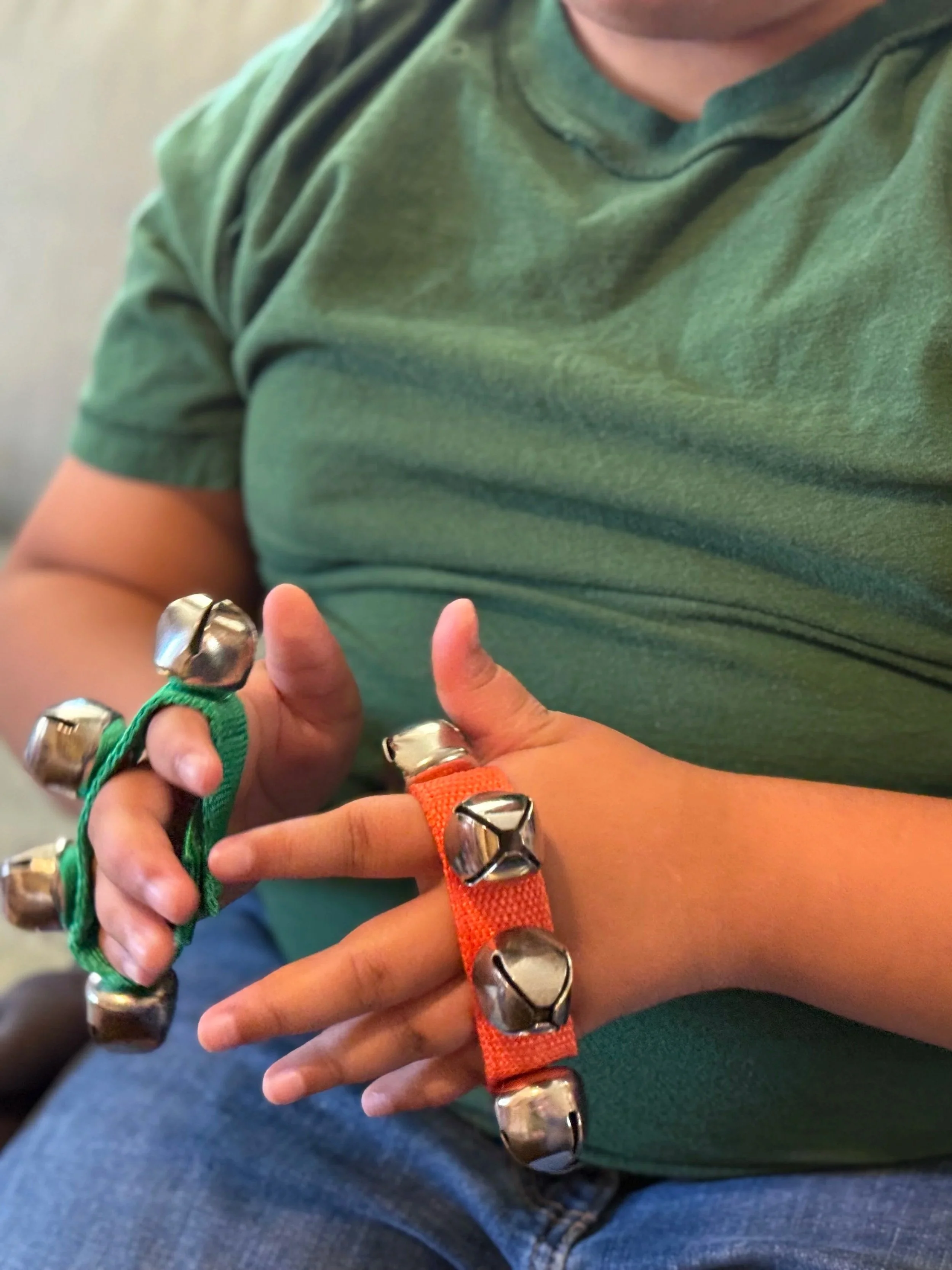 The hands of a child wearing a green shirt play with velcro jingle bell straps. One strap is green and one is orange.