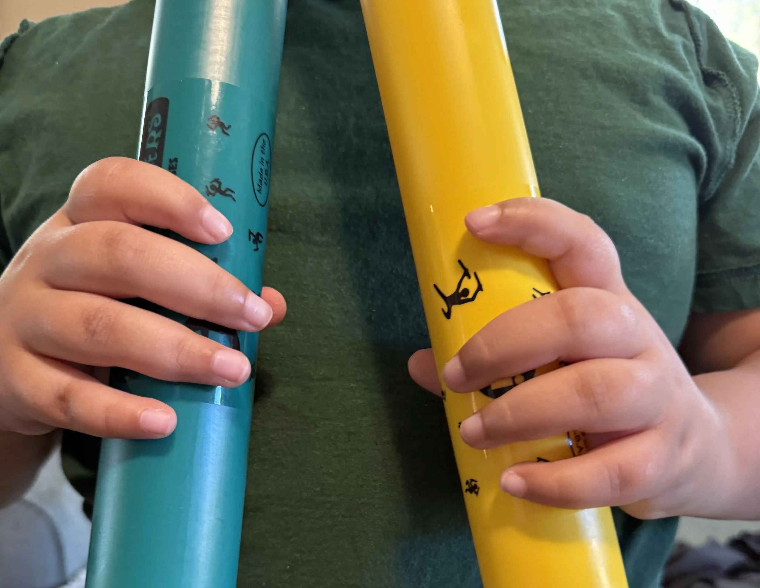 The hands of a child wearing a green shirt hold dark green and yellow boomwhackers, also known as plastic sound tubes.