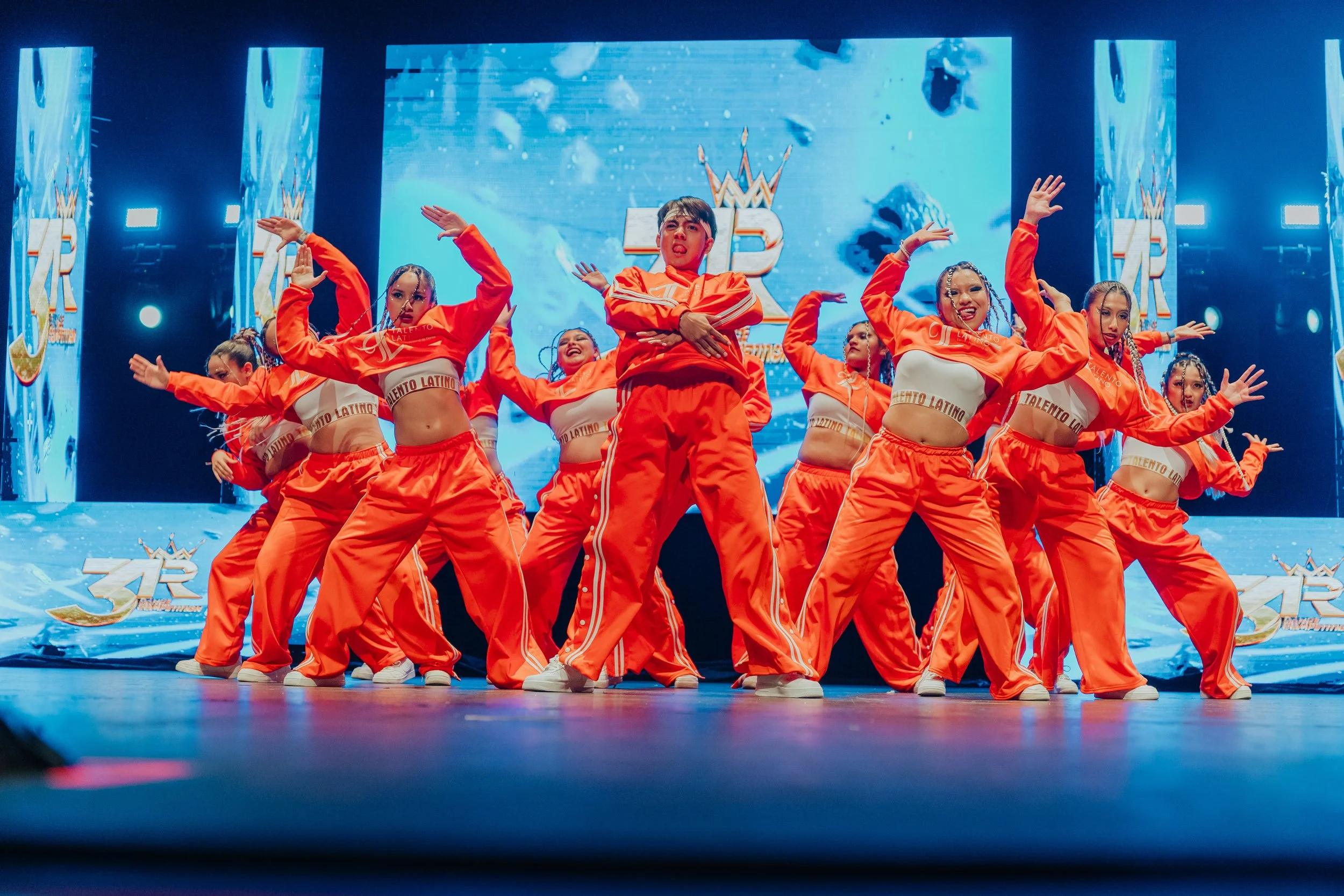 Grupo de bailarinas en un escenario, vestidas con ropa deportiva naranja y blancas, bailando en una presentación con fondo digital y logotipo de talento latino.